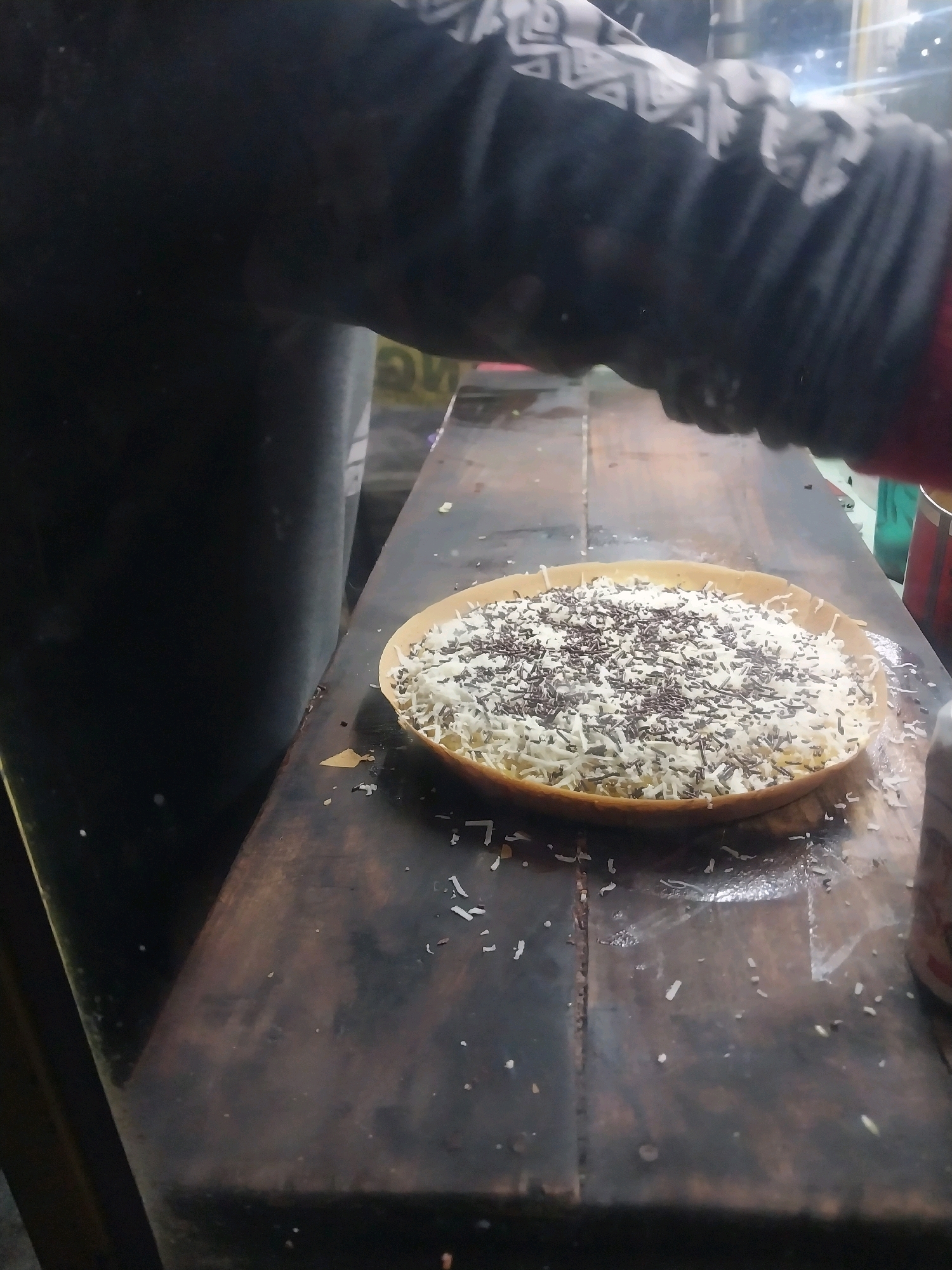 A close-up, overhead shot captures a martabak, a thick, sweet Indonesian pancake, being prepared. The martabak is filled with a generous amount of shredded white cheese and dark chocolate sprinkles, both evenly distributed across its surface. It rests on a dark, wooden surface, likely a food stall counter in the city of Nglegok, Indonesia. The surface is littered with small white flecks, likely fallen cheese. A portion of a person's arm and sleeve, clad in a dark, long-sleeved shirt with a patterned cuff, is visible at the top of the frame, suggesting they are in the process of preparing or serving the martabak. The lighting is dim, with some bright reflections visible on the wooden surface, hinting at an indoor or covered outdoor setting. The overall impression is a detailed, candid glimpse into the making of a popular street food delicacy.