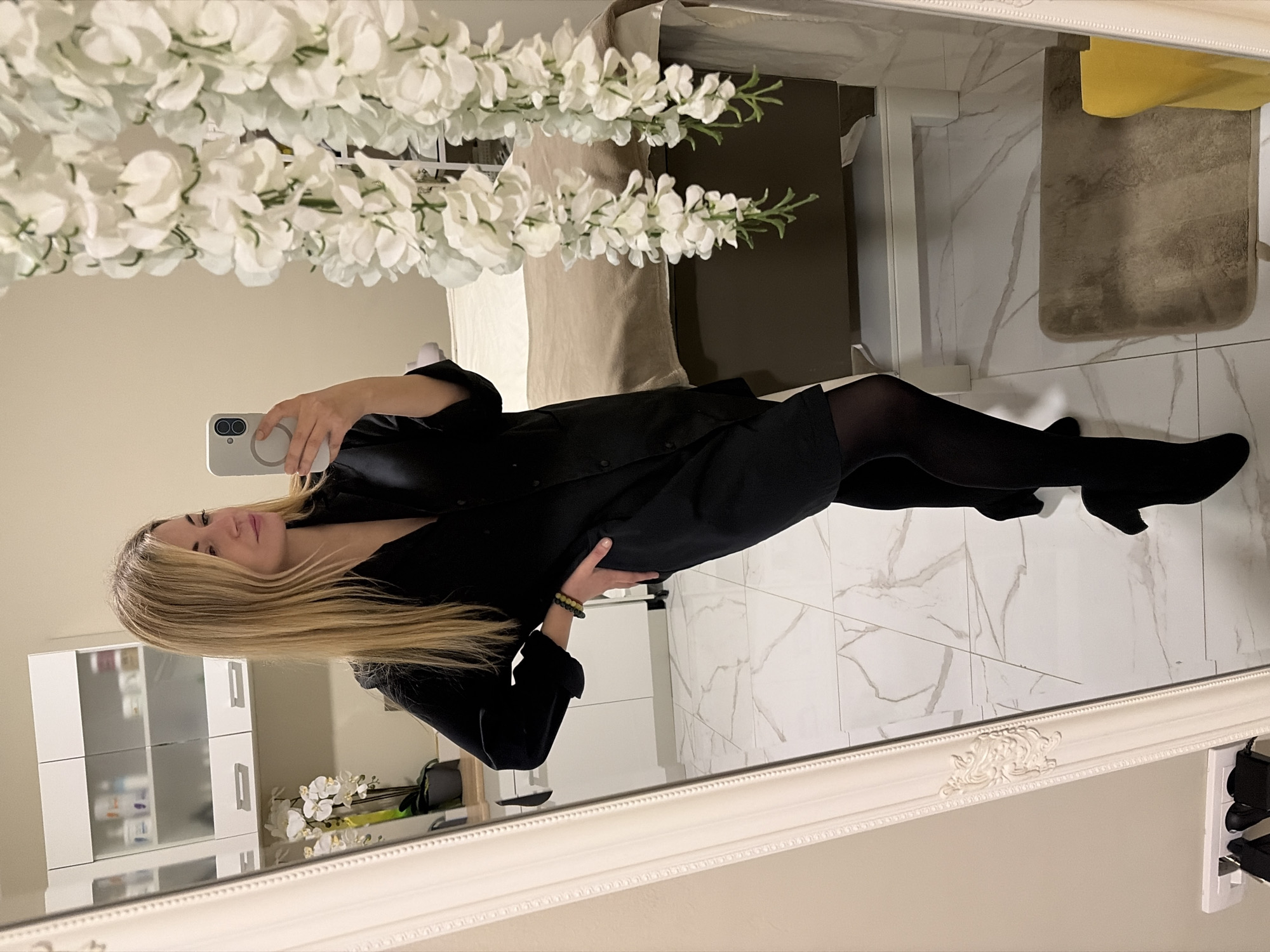 A young woman is posing in front of a large, ornately framed mirror. She is wearing a black dress and black tights. She is holding a cell phone in her right hand, presumably to take a photo of herself. Her left hand is on her hip. She has long, blonde hair. The room appears to be a bedroom. There is a bed in the background and a white cabinet with drawers. There are white orchids in a vase on a table. The floor is made of white marble tiles. A white floral arrangement is in the upper left corner of the mirror's reflection.
It is not possible to determine the specific location in Budva, Montenegro, without additional information or context.