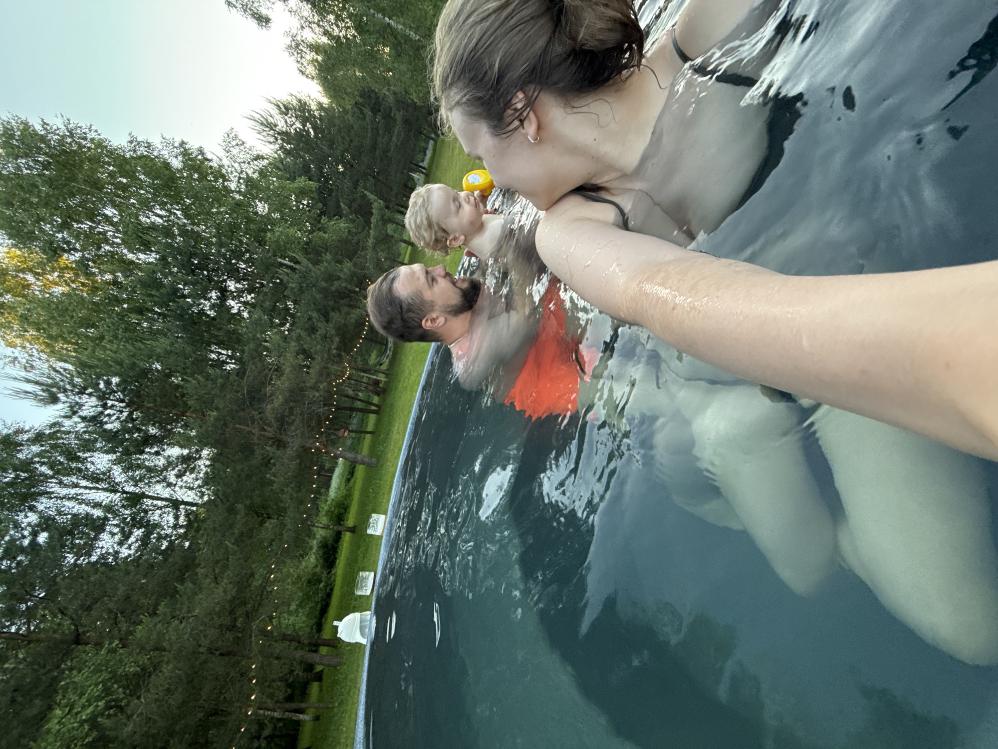 Three individuals, an adult female, an adult male, and a toddler, are depicted in a swimming pool during late afternoon or early evening in a wooded area identified as Копнино, Russia. The adult female, positioned closest to the camera on the right, has her right arm extended, indicative of taking a selfie, and is looking towards the man and child. She has wet, dark hair pulled back and wears a dark swim top. The adult male, with a beard and dark hair, is in the mid-ground on the left, submerged to his chest, wearing orange swim shorts, and is smiling while looking at the child. The toddler, with light, curly hair, is between the adults, submerged to the chest, holding a yellow plastic cup or toy, and appears to be looking towards the woman. The dark greenish-blue pool water is agitated, and the woman's submerged legs are visible. The background features dense green foliage from various trees, with multiple strands of illuminated, warm-toned string lights strung between them, suggesting an outdoor recreational setting. Green grass surrounds the pool, with a white plastic object and two clear rectangular objects resting near its edge.