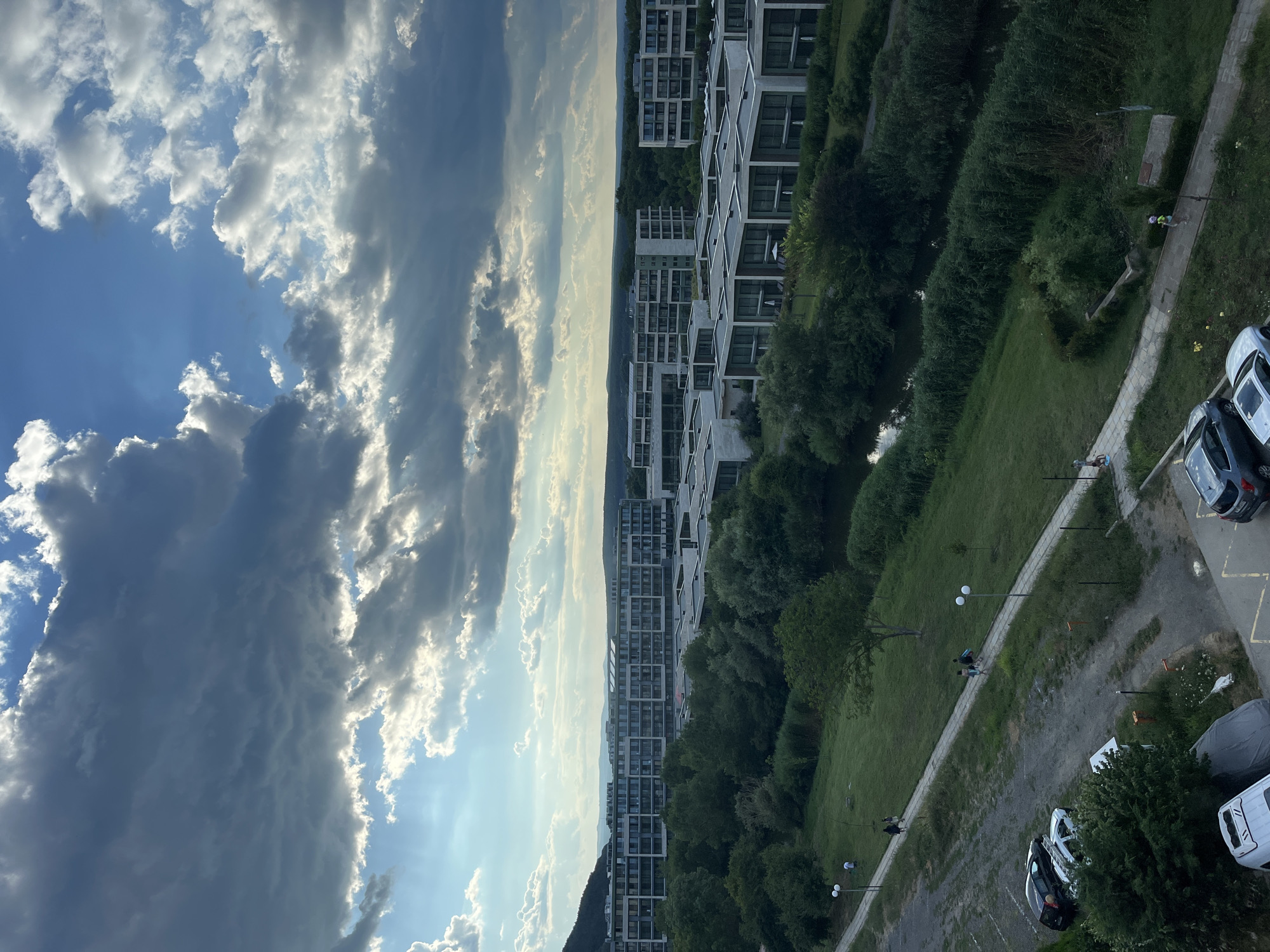 The image shows a view of modern residential buildings with flat roofs and large windows, set against a backdrop of lush green trees and a cloudy sky. The buildings appear to be part of a planned community, with pathways and a small stream winding through the landscape. Several cars are parked in a gravel lot in the foreground, and a few people are visible walking along the pathways. The scene suggests a contemporary residential area possibly located on the outskirts of a city or town, potentially in a scenic natural setting. The lighting indicates it might be late afternoon or early evening, with sunlight breaking through dramatic clouds. The location is identified as Obzor, Bulgaria.
