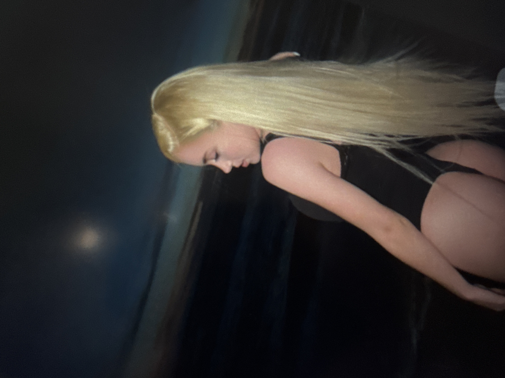 An image, rotated on its side, features a person with very long, straight blonde hair fanned out to the right. The individual is positioned with their head tilted downwards and to the left, revealing a profile view of their face. They appear to be wearing a dark, possibly black, one-piece garment such as a leotard or swimsuit. Their skin tone is light.
The setting is dark and indistinct, suggesting low-light conditions, possibly indoors or at night. In the upper left background, a soft, circular light source is faintly visible. The overall background is a deep blue-grey to black, with some subtle vertical lines that could indicate a textured wall or curtains. The image primarily focuses on the person, highlighting the contrast between their bright blonde hair and the dark surroundings. No specific emotions are discernible, and no text is visible.