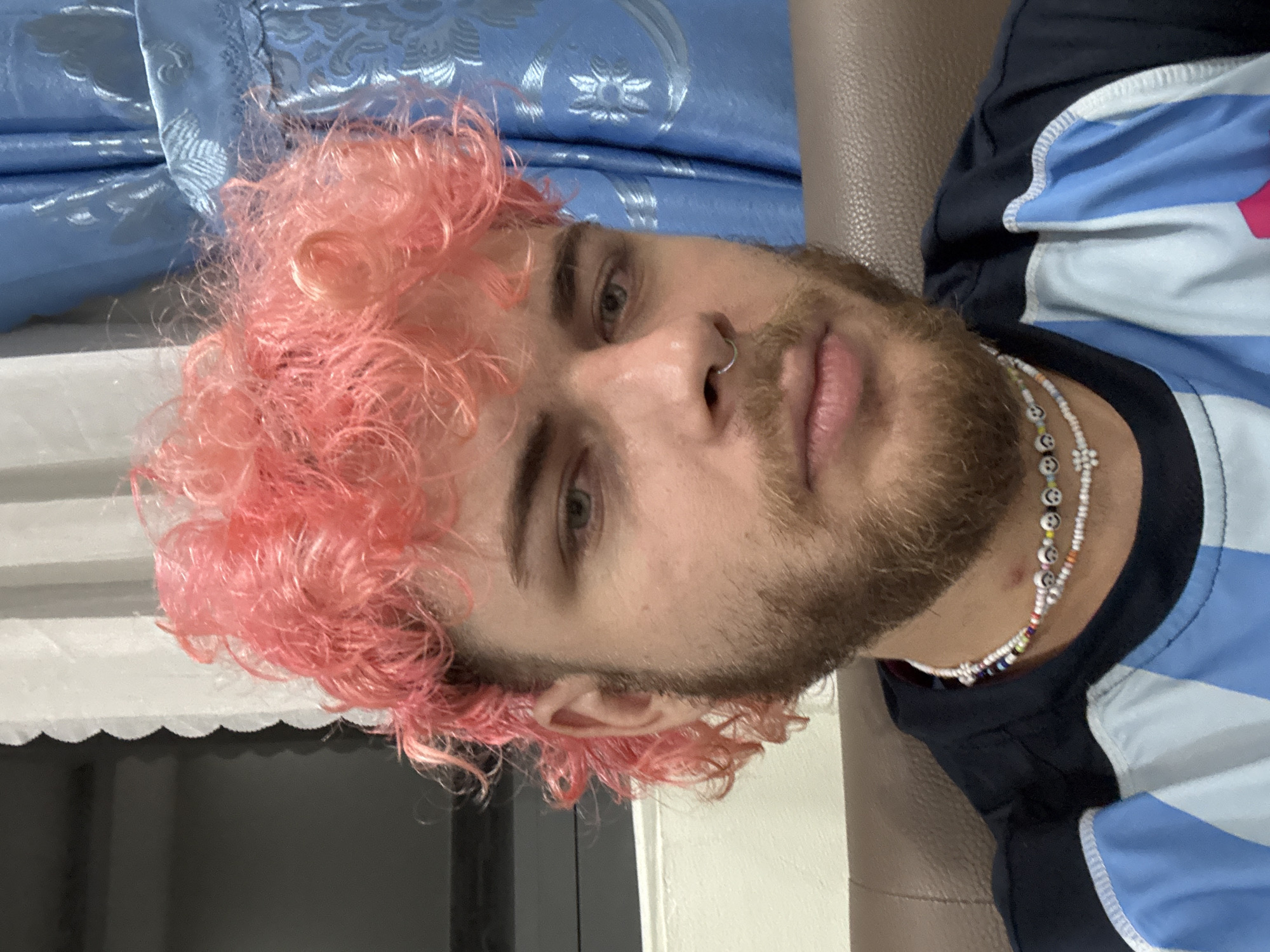 This is an indoor portrait of a young adult male in Đà Nẵng, Vietnam. The subject, positioned centrally, looks directly at the camera with a neutral expression. He has light skin, light-colored eyes (appearing blue or green), and light brown facial hair comprising a beard and mustache. His hair is dyed a vivid pink, styled with short, curly locks on top and longer, curly sections at the sides and back, consistent with a modern mullet haircut. A silver hoop septum piercing is visible in his nose.
He is wearing a dark blue athletic-style shirt with prominent light blue and white striped sections on the chest and shoulders. Around his neck, two beaded necklaces are visible: one features small, colorful beads interspersed with black and white "smiley face" beads, and the other, a shorter choker, is composed mostly of white beads with a central white floral bead and smaller dark accents.
The background elements are out of focus. To the subject's left, a white, possibly ruffled or scalloped, vertical border is visible against a dark grey background. To his right, a section of a textured, light blue curtain with a prominent repeating pattern, possibly floral or damask, is present. The subject appears to be seated on a piece of furniture upholstered in a dark brown, leather-like material, visible behind his right shoulder and in the lower right corner of the frame.