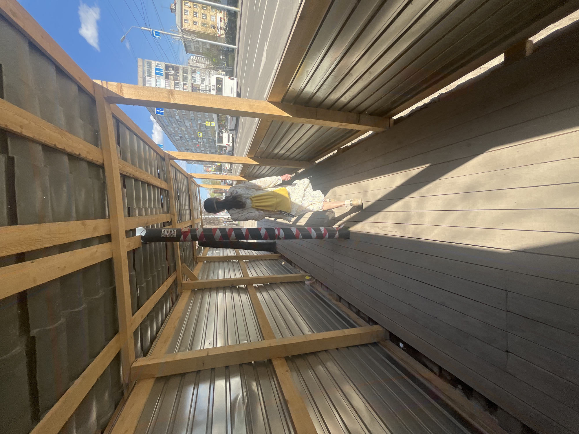 Here is a detailed description of the image:
**Scene:** An outdoor setting, possibly a covered walkway or passage under construction, in a city. The perspective is slightly rotated clockwise.
**Location:** The location is in an urban environment of unknown city and state. Buildings are visible in the background across a wide roadway.
**People:** A person wearing a light-colored patterned dress with a yellow underdress and black cap, is lying on their back on a wooden surface.
**Objects:**
* **Construction Structure:** A framework of wooden beams supports a corrugated metal roof, forming a covered walkway.
* **Piping:** Secured along the metal walls are long, black pipes with red and white striped bands.
* **Wooden Surface:** The floor of the structure is made of planks, probably wood.
* **Street:** Beyond the walkway, there is a street with cars and buildings.
**Actions:** The person is lying down, seemingly in a relaxed or casual posture.
**Interactions:** The person is interacting with the built environment, likely resting or posing for a photograph.
**Environment:** It is a bright, sunny day. The city in the background appears densely populated.
