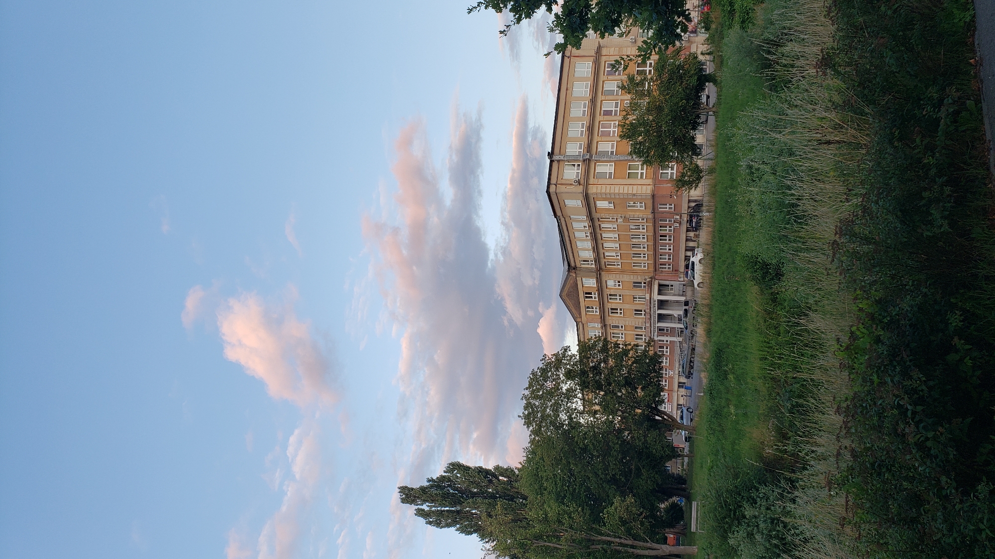 The image depicts a large, multi-story building with a beige facade, featuring numerous windows and a symmetrical architectural design. The building appears to be a school or institutional facility, possibly the Faculty of Arts of the University of Prešov in Prešov, Slovakia, based on the context of similar structures in the city. The building is situated behind a wide expanse of green grass and vegetation, with several trees in the foreground. The sky above is a clear blue with scattered, wispy clouds tinged with pink and orange, suggesting it is either sunrise or sunset. Several cars are parked in front of the building, and a bench is visible to the left of the building. The overall scene is calm and serene, capturing a moment of quietude in an urban environment.