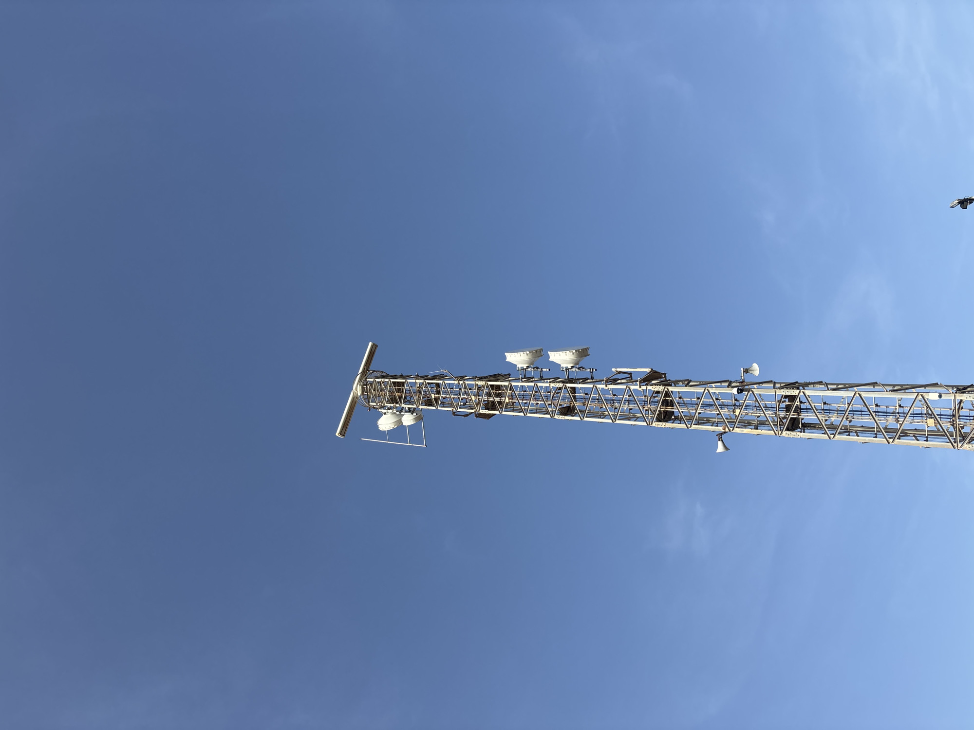 The image shows a tall, metal telecommunications tower against a clear blue sky. The tower is a lattice structure with several antennas and dishes attached at various levels. A radar-like device is mounted at the very top. The tower appears to be located in an open area, as there are no other structures or significant vegetation visible in the immediate vicinity. The image does not provide enough information to determine a specific location within the city of Orihuela, Spain, or to identify any people or specific actions.