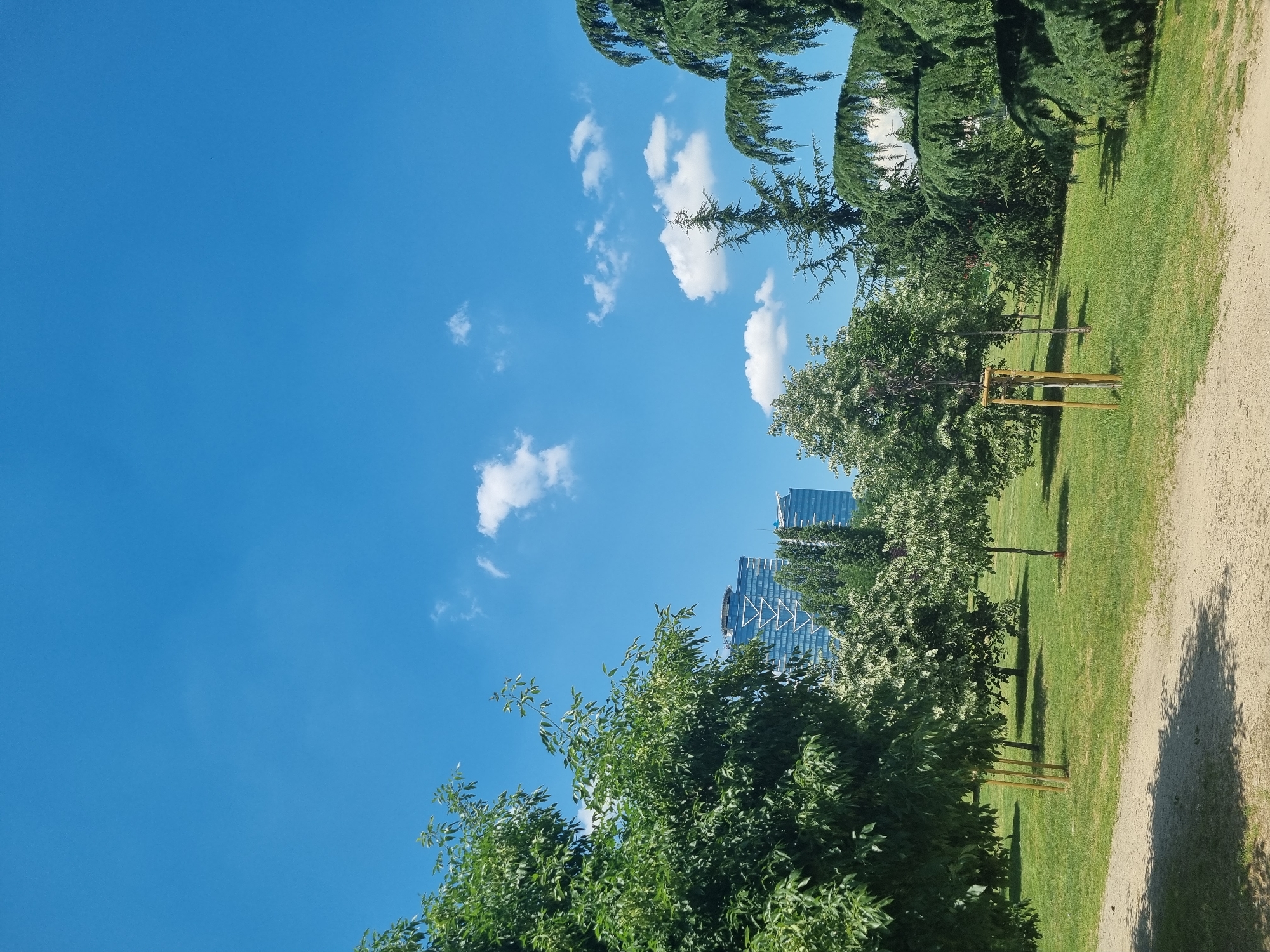 The image depicts a park scene with lush green grass, various trees, and a clear blue sky with scattered white clouds. In the background, modern high-rise buildings are visible. The park appears to be well-maintained with a dirt path in the foreground casting shadows from the trees. The presence of young trees staked with wooden poles suggests recent planting or landscaping. The architectural style of the buildings in the background indicates an urban setting. Based on the distinctive architectural features of the buildings, this scene is likely located in the Business Park Sofia, a prominent business district in Sofia, Bulgaria. There are no people visible in the image.