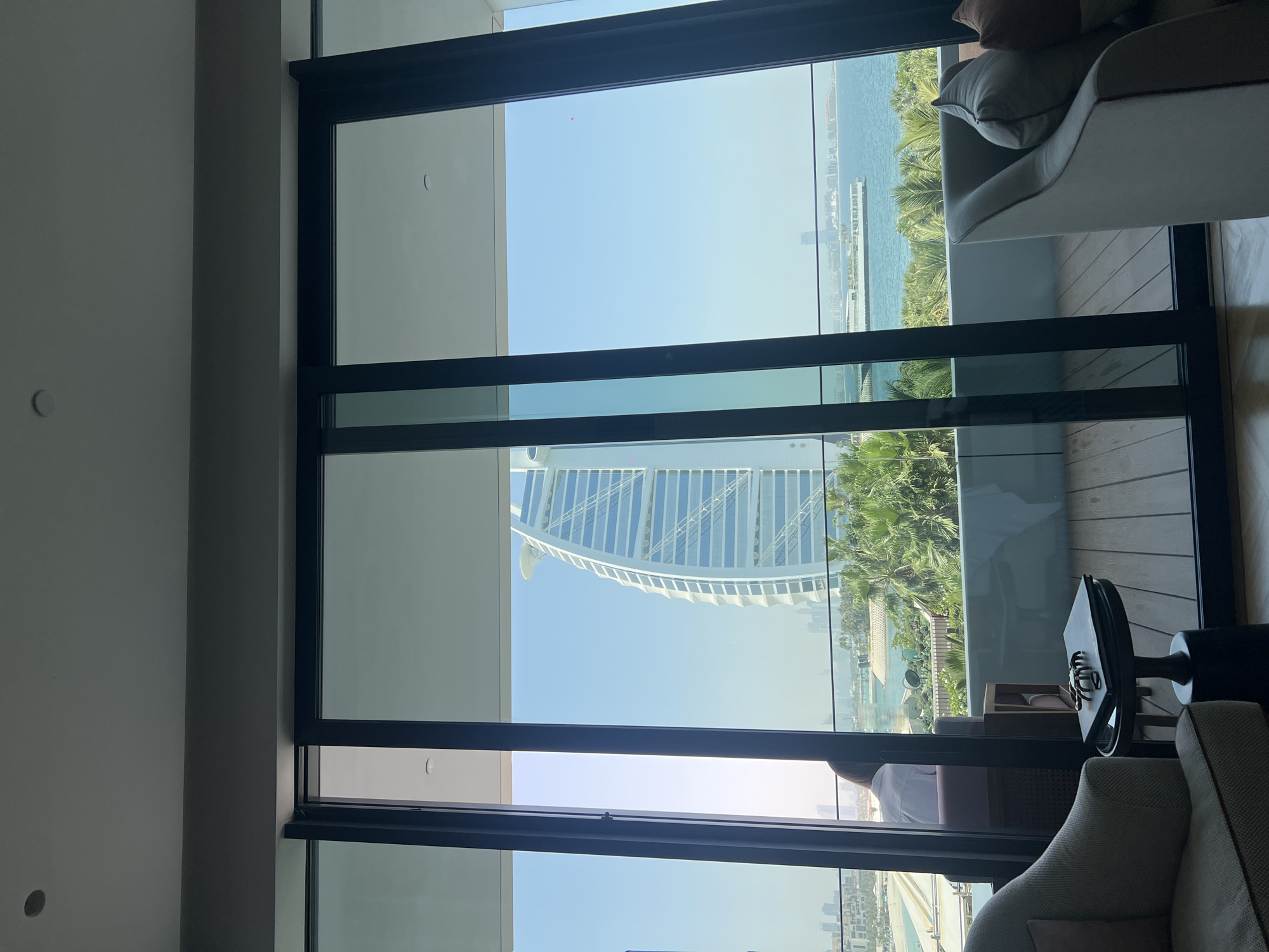 The image depicts an interior view through a large window showing the Burj Al Arab, a luxury hotel in Dubai, United Arab Emirates. The window, framed in dark metal, is divided into panes. Outside, palm trees are visible between the building and the sea, with other buildings in the distance. Inside, part of a gray sofa with a brown pillow is visible on the right. There is also a black side table with items on it. The interior walls are painted in a light neutral color.