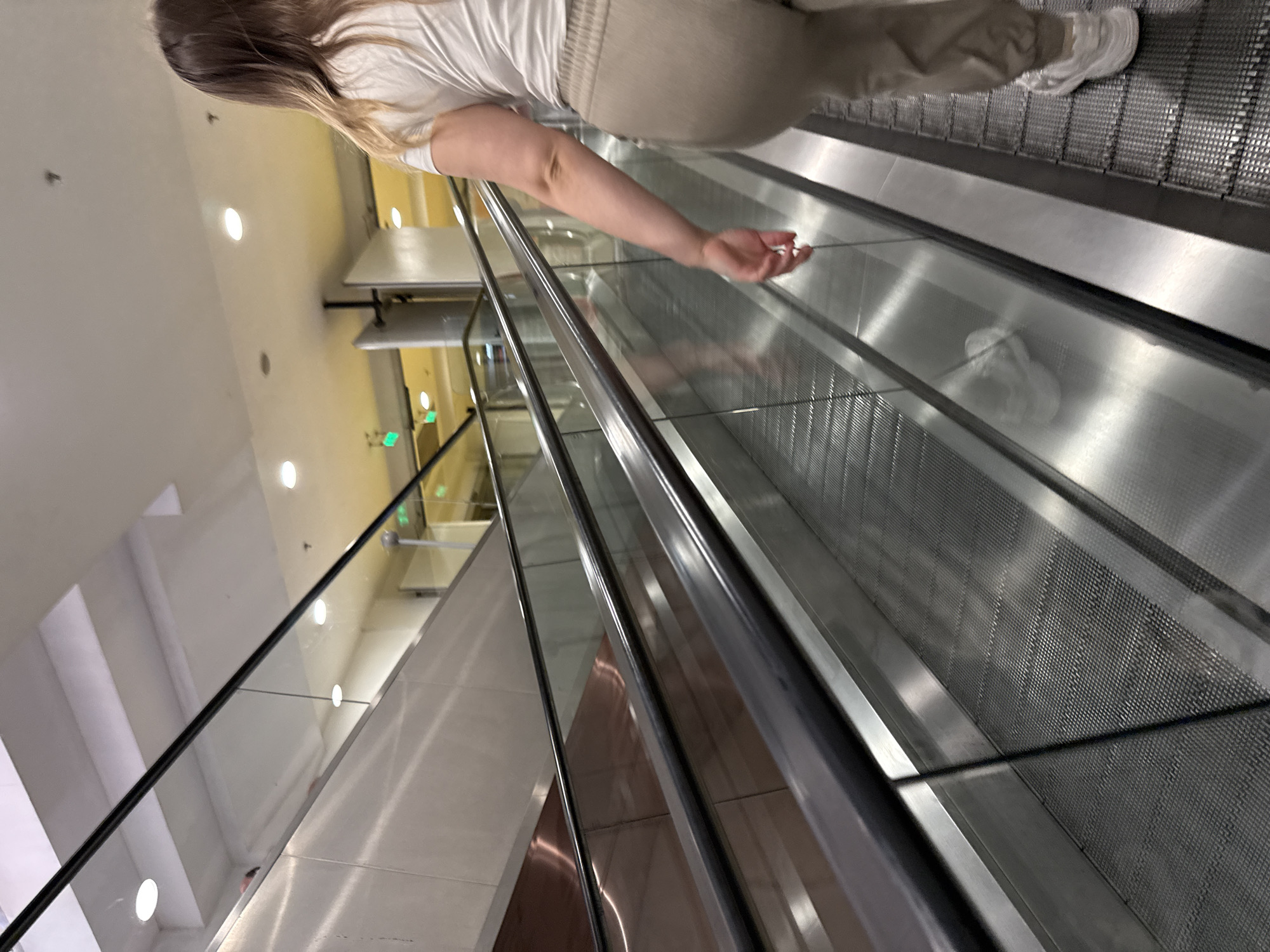 A woman is standing on a moving walkway. She is wearing a white shirt and beige pants. The walkway is located in an indoor setting, possibly an airport or shopping mall, with a view of the building's architecture. The location details are not discernible.