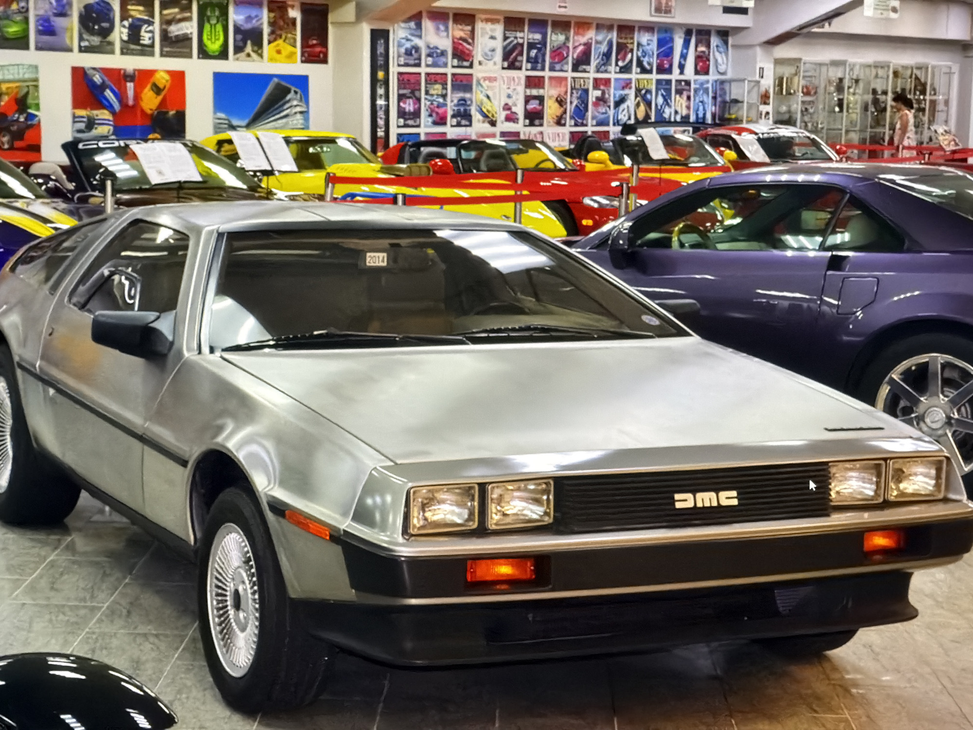The image shows a silver DeLorean car in what appears to be a car museum or showroom, with other cars visible in the background. The DeLorean is in the foreground and angled slightly to the left. Behind it, a purple car is partially visible. Further back, there are yellow and red cars, along with a display of automotive-themed posters or photographs on the wall. In the upper right of the image, there is a person standing near display cases. The location is in the city of Unknown, Unknown.