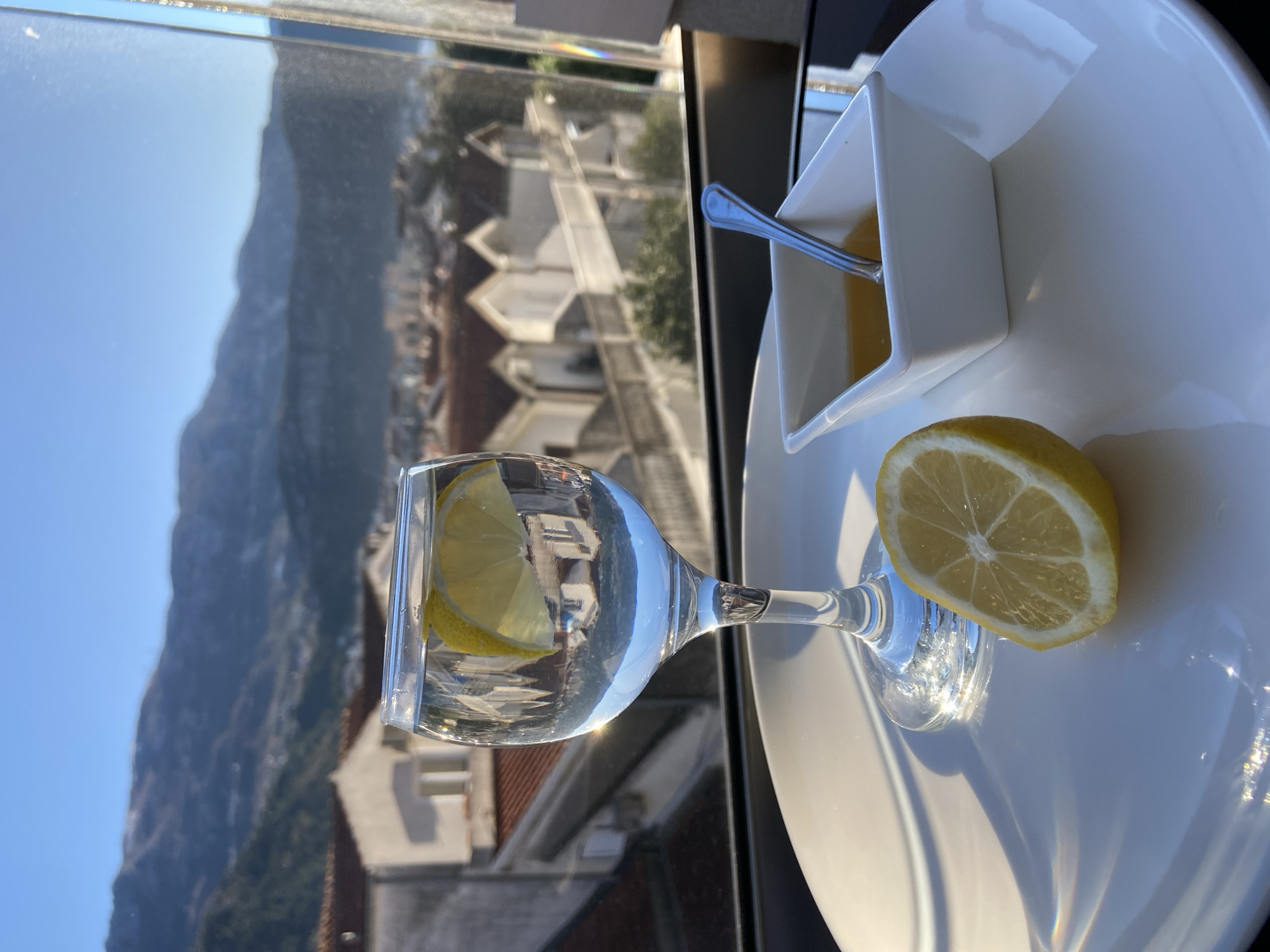 A glass of water with a lemon slice inside sits on a white plate, next to a lemon slice and a square dish filled with a yellowish liquid and a small spoon. The plate rests on a dark surface, possibly a table. Through a window, a view of Budva, Montenegro can be seen, including buildings with red tile roofs and a large mountain in the background. The sky is clear and blue. The scene is well-lit, suggesting it is daytime.