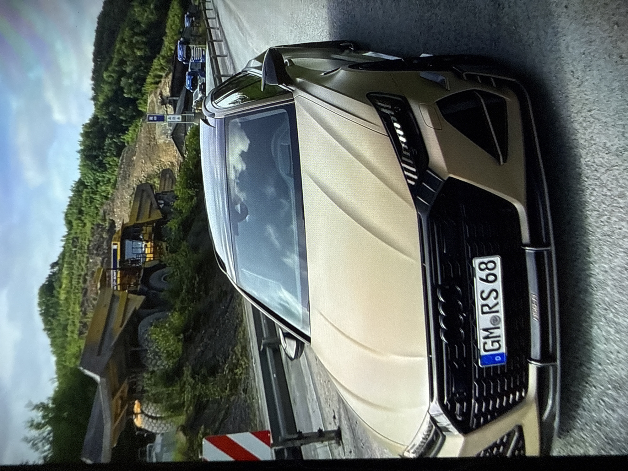 This image depicts an outdoor daytime scene in the city of Varna, Bulgaria. A matte gold Audi RS6 Avant, bearing a German license plate "GM RS 68", is positioned in the foreground, slightly angled towards the camera from its front-left. The vehicle features a large black honeycomb grille with the Audi emblem, illuminated LED headlights, and dark roof rails. The road surface beneath the car is asphalt, and a red and white striped warning marker is visible on the far left.
In the mid-ground, on the left, a large-scale excavation or mining site is visible, characterized by elevated, dusty terrain and sparse vegetation. Two massive yellow-orange mining dump trucks are present; the one closer to the foreground has its bed raised. Dense green trees cover the hills behind this industrial area.
On the right side of the mid-ground and extending into the background, a multi-lane highway or major road is visible, bordered by a metal guardrail. Several blue-cab semi-trailer trucks are parked or traveling in the distance along this road. Structures and a tall blue and white sign, possibly indicating a service station or rest area, are discernible near the highway. The sky above is overcast with scattered clouds, providing diffused natural lighting to the scene. No individuals are visibly present in the image.