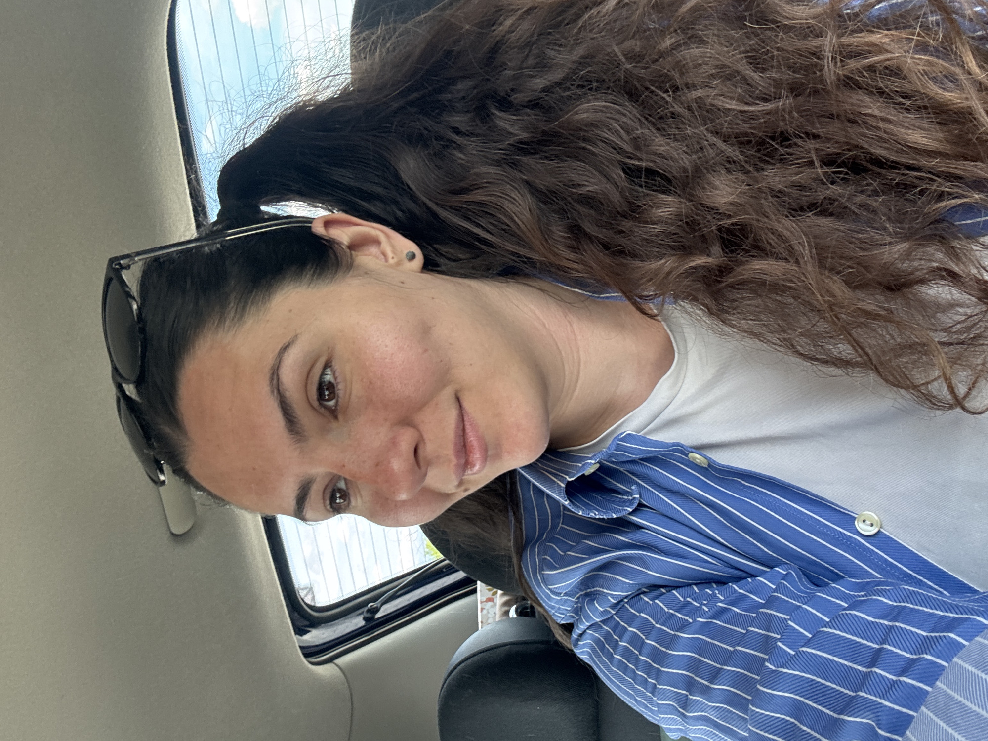 The image depicts a woman with brown hair, partially pulled back, wearing sunglasses on her head. She has brown eyes and is wearing a white t-shirt under a blue and white striped button-down shirt. She is sitting in a car. The location is listed as Unknown, Unknown.