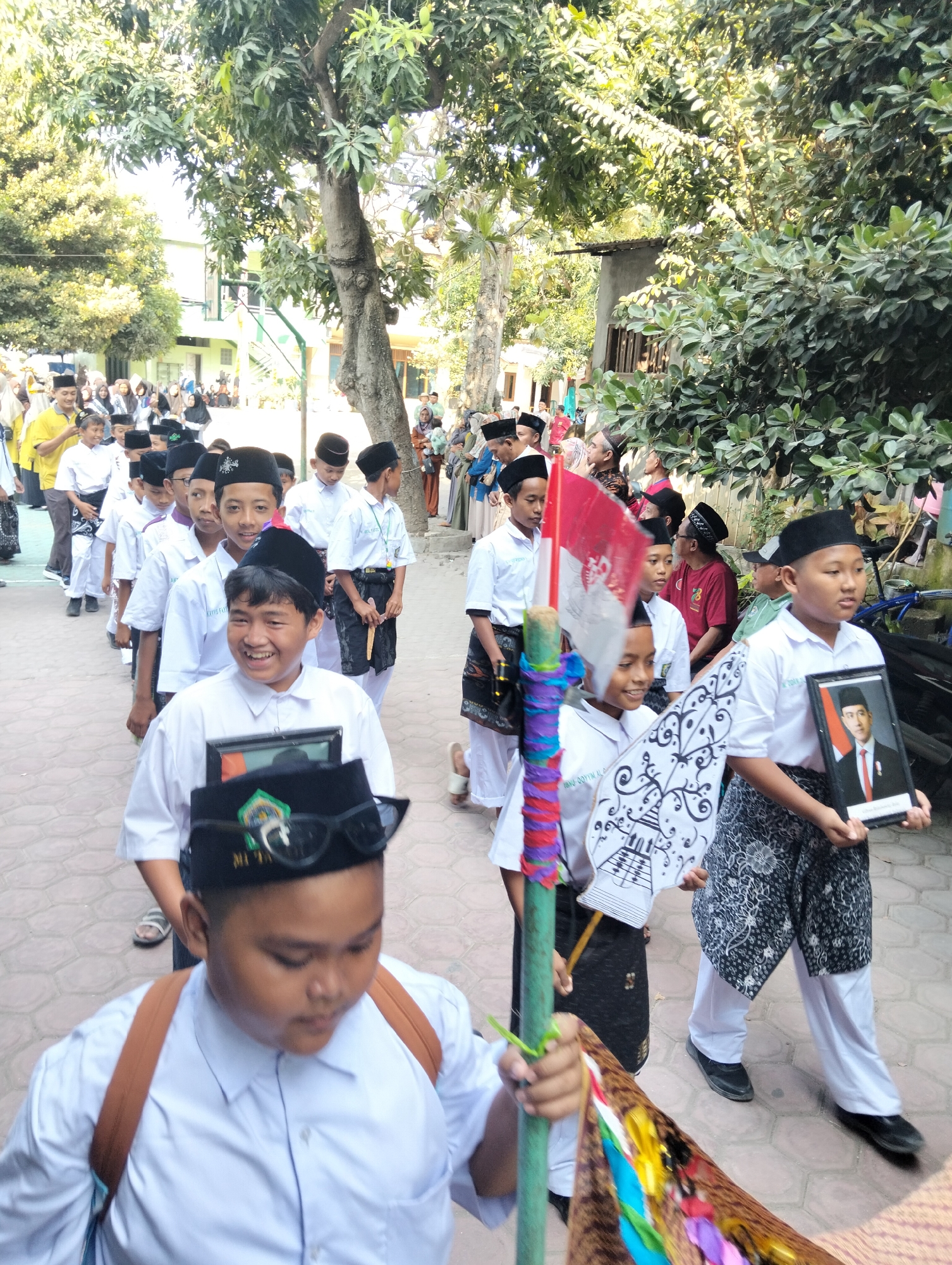 This is a vertical, medium-angle, outdoor shot of a procession of young boys in white shirts and black caps, walking on a paved path lined with trees. The procession appears to be part of a school event in Kranji, Indonesia.

In the foreground, a boy with a black cap adorned with sunglasses and an emblem is smiling and looking down. He is wearing a white shirt and a brown backpack. Behind him, a line of boys, also in white shirts and black caps, continues down the path. Some are holding flags, one of which is the Indonesian flag. Another boy is holding a framed photograph of a man in a suit.

In the background, more people, including adults and children, are visible. They are gathered on the sides of the path and in front of buildings, some of which appear to be school structures. The environment is characterized by lush green trees and dappled sunlight, suggesting a warm, sunny day. The overall atmosphere appears festive and orderly.
