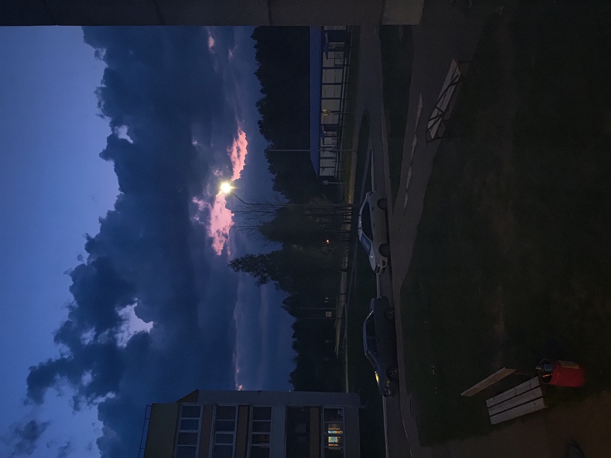 The image depicts an urban scene at dusk in Марковский, Russia. The sky is a deep blue, with dark clouds illuminated by a street lamp creating streaks of pink. The scene includes residential buildings, trees, parked cars, benches, a section of sidewalk, and a red object possibly a container. The overall environment suggests a quiet, residential area at twilight.