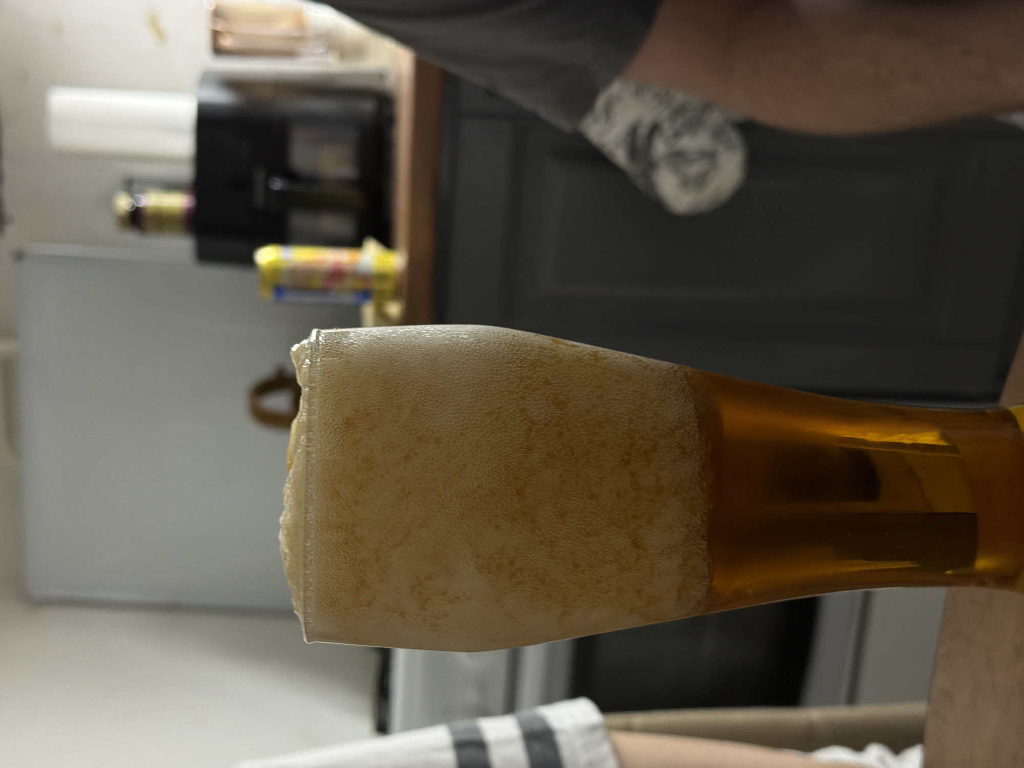 A tilted close-up shot shows a partially-full glass of beer with a large foamy head. The beer is golden in color. A blurry background includes kitchen cabinets, a refrigerator, and various objects on a countertop. A person's arm and leg are partially visible in the foreground and background. Location is not discernible from the image, and Reshetnikovo, Russia, cannot be determined.