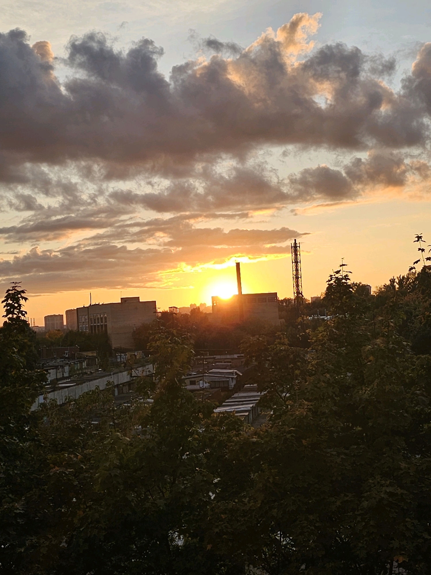 This image depicts a sunset over a city landscape in Khimki, Russia. The sky is filled with dramatic clouds, tinged with orange and yellow hues from the setting sun. The sun itself is partially obscured by clouds, casting a bright glow over the scene. In the foreground, lush green trees with dark foliage dominate the lower part of the frame, with some appearing to be silhouetted against the brighter sky. Behind the trees, a cityscape unfolds. There are several buildings, some appearing to be industrial or utilitarian in design, with flat roofs. One prominent structure is a tall, thin tower with a lattice-like appearance, possibly an antenna or industrial chimney, located to the right of the setting sun. Below the main buildings, there are rows of what appear to be garages or small storage units. The overall impression is one of an urban environment at the end of the day, with a mix of industrial and residential elements visible under a vibrant sky. There are no people or discernible actions visible in the image.