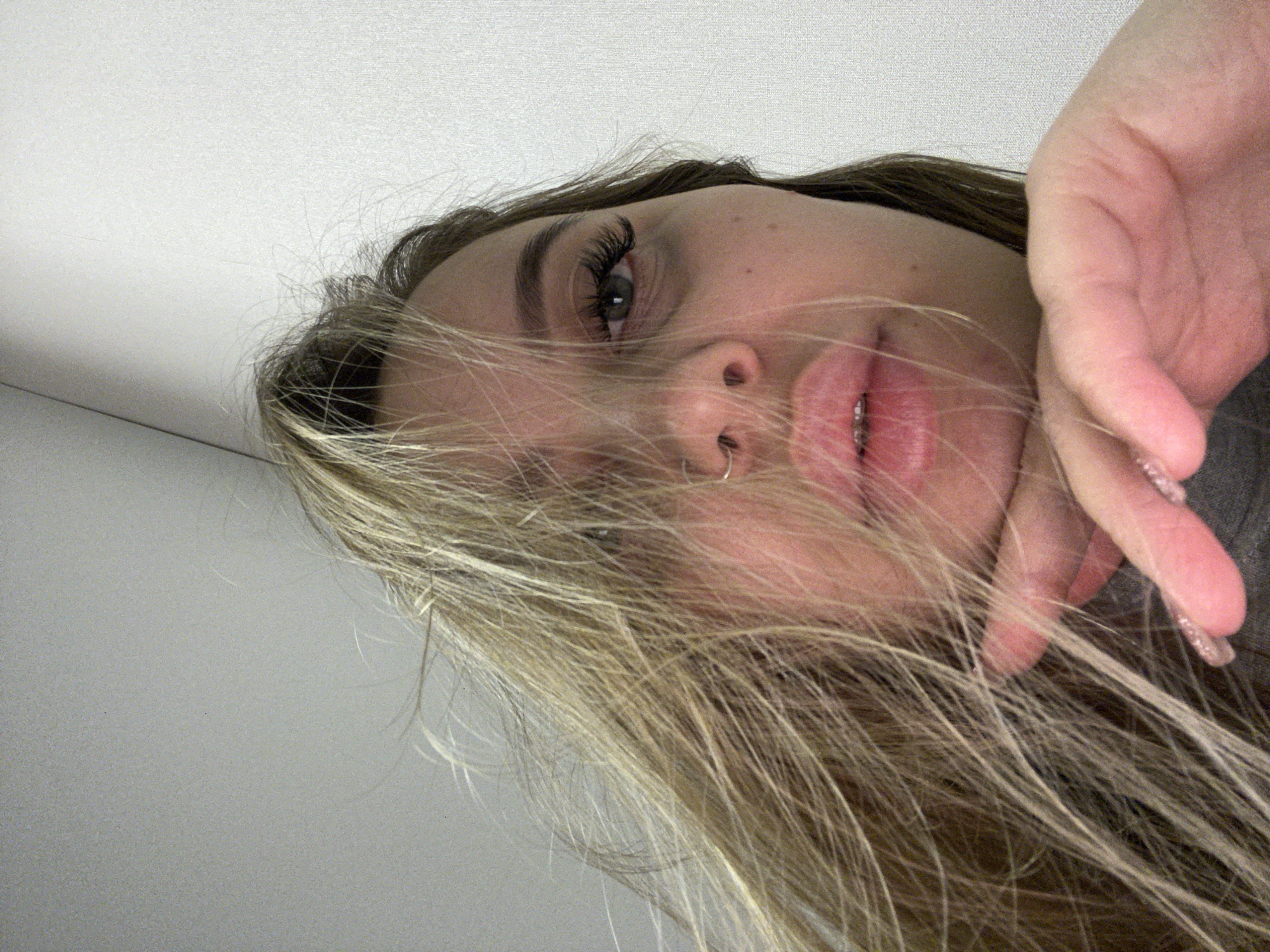 A selfie taken from a low angle shows a person with long, blonde hair partially obscuring their face. The person has a nose ring and appears to be wearing eyelash extensions. Their hand is raised near their face. The background is a plain, light-colored wall. The location is not discernible from the image.