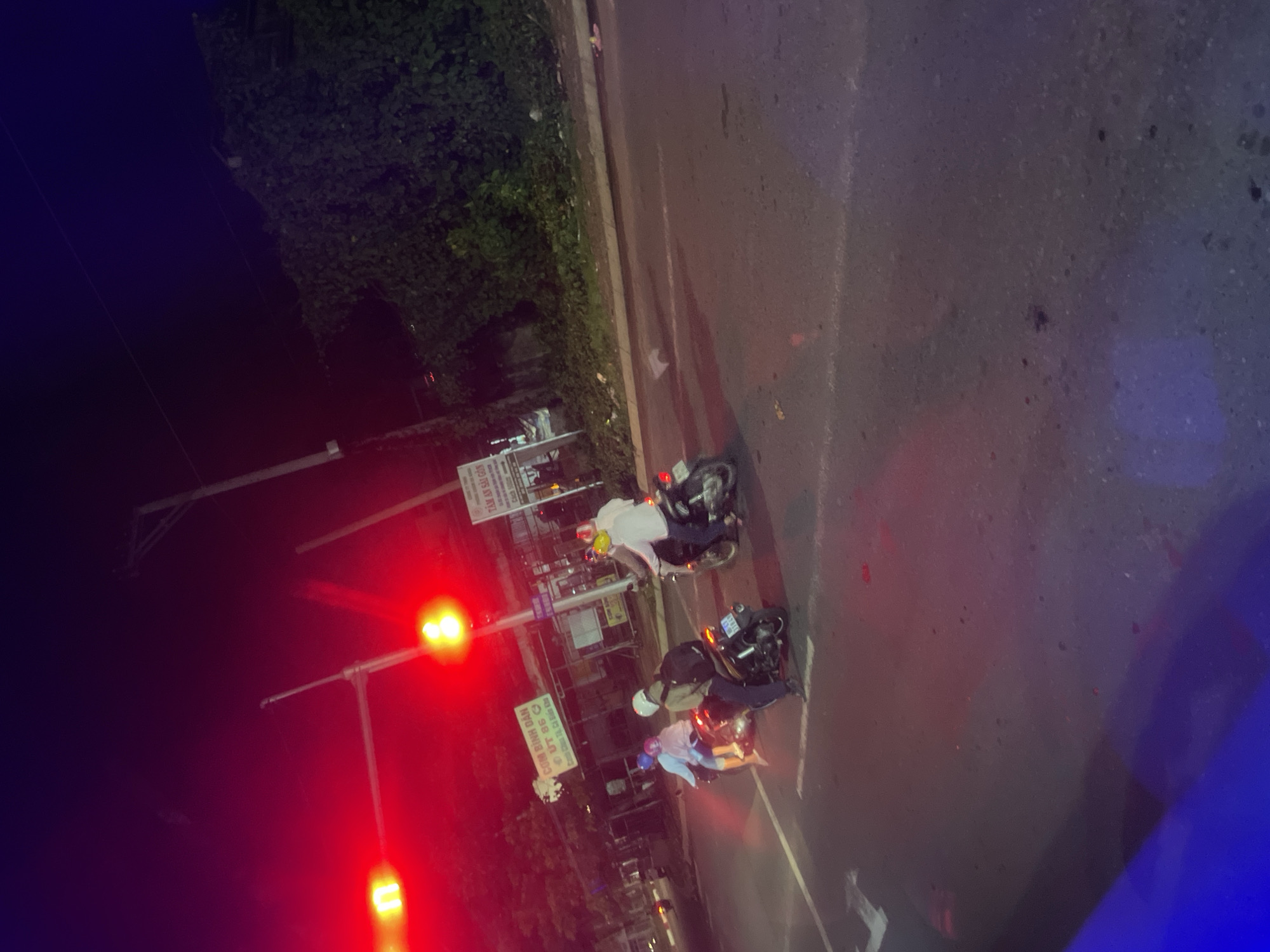 The image shows a nighttime scene on a street in Binh Thuan Ward, Vietnam. Several motorbikes are present, with people riding them. One motorbike in the foreground has a license plate that reads "61F1-181.41". Another motorbike has a rider wearing a yellow helmet and a white long-sleeved shirt. There are also people on a motorbike with a blue helmet and another with a beige helmet and a backpack. Traffic lights are visible, illuminated by red and yellow lights. A sign with Vietnamese text, possibly advertising food, is mounted on a pole. The street appears to be paved, with white lane markings. Vegetation, likely bushes or trees, is visible in the background, and there is a street light pole. The overall impression is of a busy street at night with motorbikes traversing it.