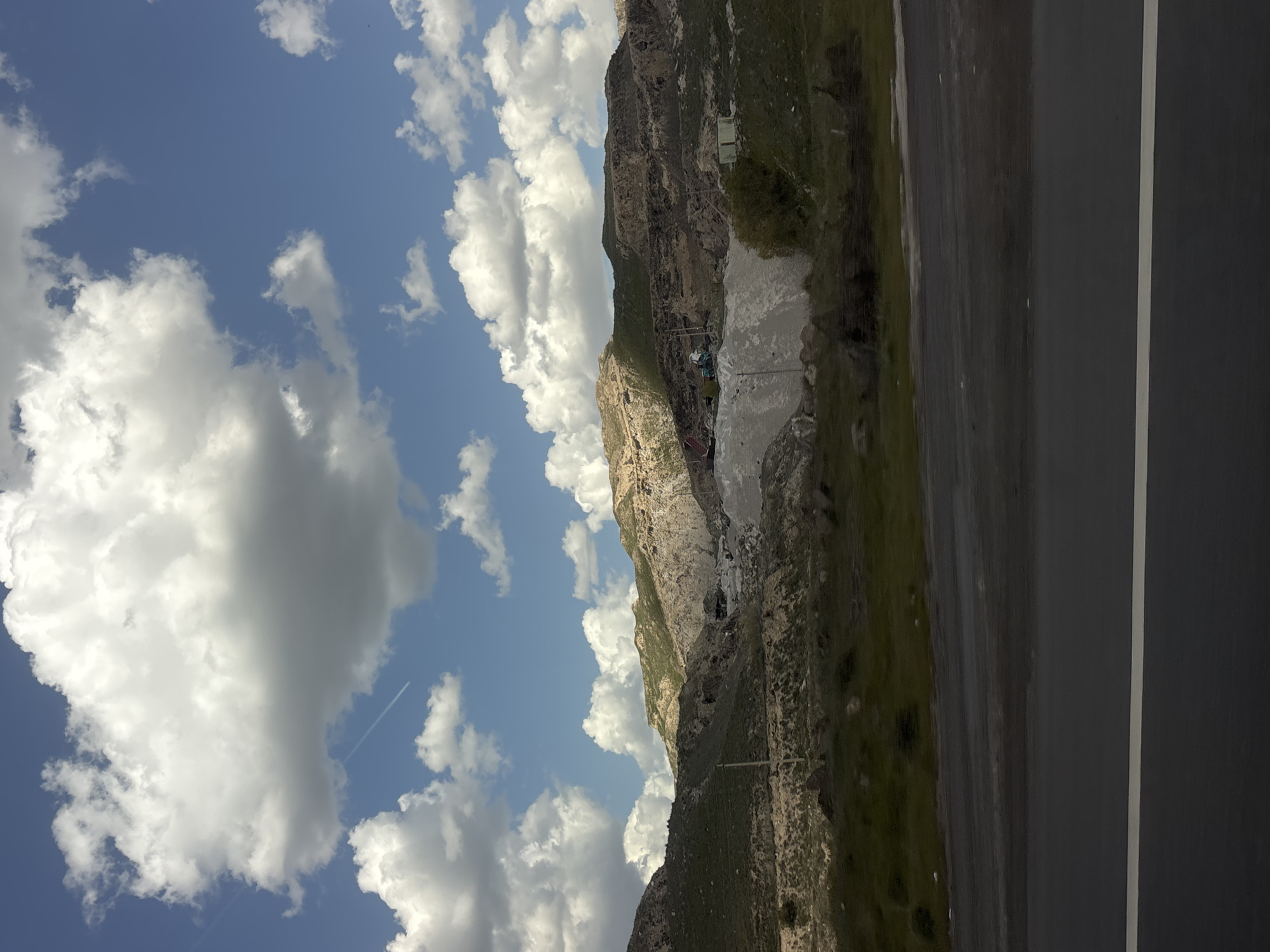 The image shows a landscape in Arzni, Armenia. A mountain with rocky terrain and sparse vegetation is visible. A road runs alongside the mountain, featuring a white line marking the lanes. The sky is blue with scattered white clouds. The scene is captured from a perspective looking slightly upwards towards the mountain and sky.