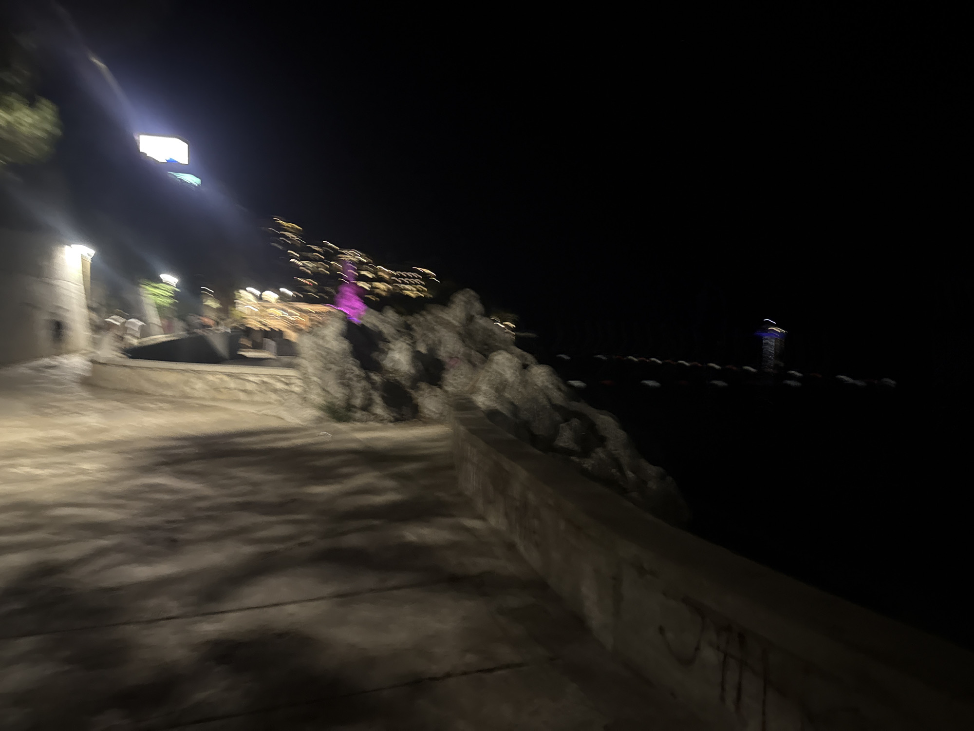 This is a nighttime photograph taken from a walkway in Budva, Montenegro. The walkway is paved with concrete and has shadows cast on it, suggesting it is illuminated by artificial lights. To the left of the walkway, there is a stone wall and some vegetation. Further to the left and above, a brightly lit square object, possibly a sign or a building facade, is visible against the dark sky.
To the right of the walkway, there is a rocky incline. Above the rocky incline and further in the distance, numerous small lights are visible, indicating buildings or a cityscape. The sea is to the far right of the image, with a faint, glowing structure in the distance that could be a lighthouse or a boat. The overall impression is of a coastal area at night with some signs of urban activity. The image is slightly blurred due to motion or low light conditions.