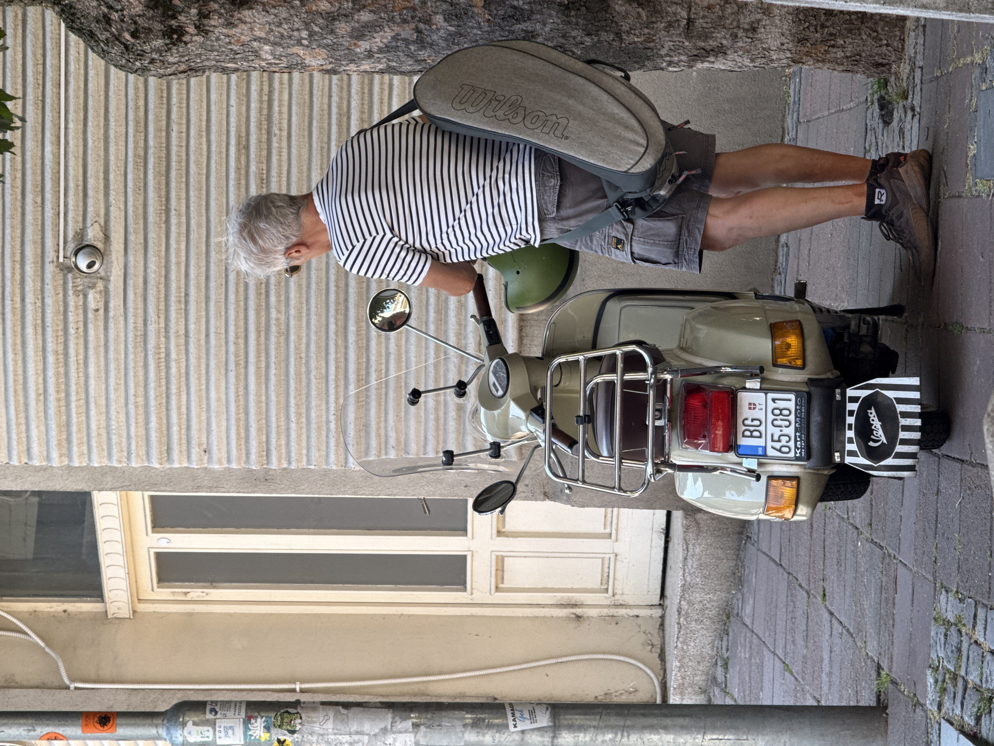A light-skinned man with graying hair, wearing a black and white striped t-shirt, gray cargo shorts, dark brown athletic shoes, and dark socks, stands with his back to the camera, facing a light-colored Vespa scooter. He holds a green helmet in his left hand and has a gray duffel bag with "Wilson" printed on it slung over his right shoulder. The Vespa scooter has a windshield, a chrome rack on the front, a brown seat, and a rear license plate that reads "BG 65-081". A black and white striped plate with the "Vespa" logo is attached to the rear fender. The scene is set on a cobblestone sidewalk next to a building with a corrugated metal exterior. A CCTV camera is mounted on the building wall. The man and scooter are positioned in front of a tree trunk. The overall impression is that the man is either about to mount or has just dismounted the scooter. The license plate and building architecture suggest the location is Belgrade, Serbia.