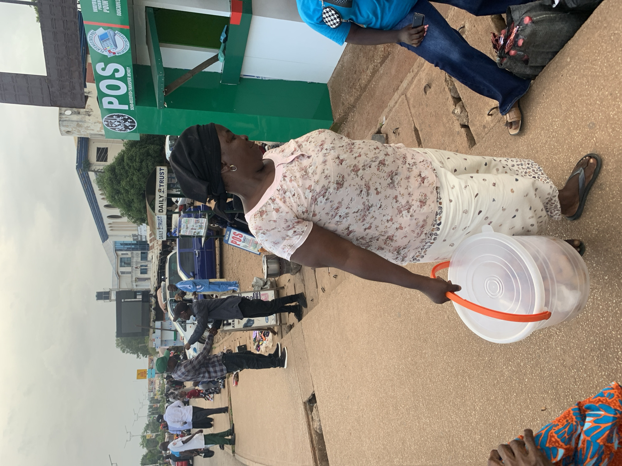 The image captures an outdoor scene in the city of Barakin Lerawi, Nigeria, on a cloudy day. In the foreground, a woman wearing a black headscarf and a floral-patterned blouse carries a large, transparent plastic bucket with orange handles. She is seen from the waist down, walking on a sandy, cracked sidewalk. To her right, another person in a blue t-shirt and jeans is partially visible, holding a smartphone.
Further back, a vibrant green POS (Point of Sale) booth is prominent, with signage indicating services for "SEND-RECEIVED-TRANSFER MONEY." People are gathered around this area. A man in a plaid shirt and a green cap appears to be interacting with another man in a dark shirt and dark pants, with what might be a transaction or a greeting taking place. Several other individuals are visible in the background, along with cars and buildings lining the street, suggesting a busy urban environment. Signs for "DAILY TRUST" are also visible, indicating a newspaper vendor or distribution point. The overall impression is of a bustling street scene with commercial activity.