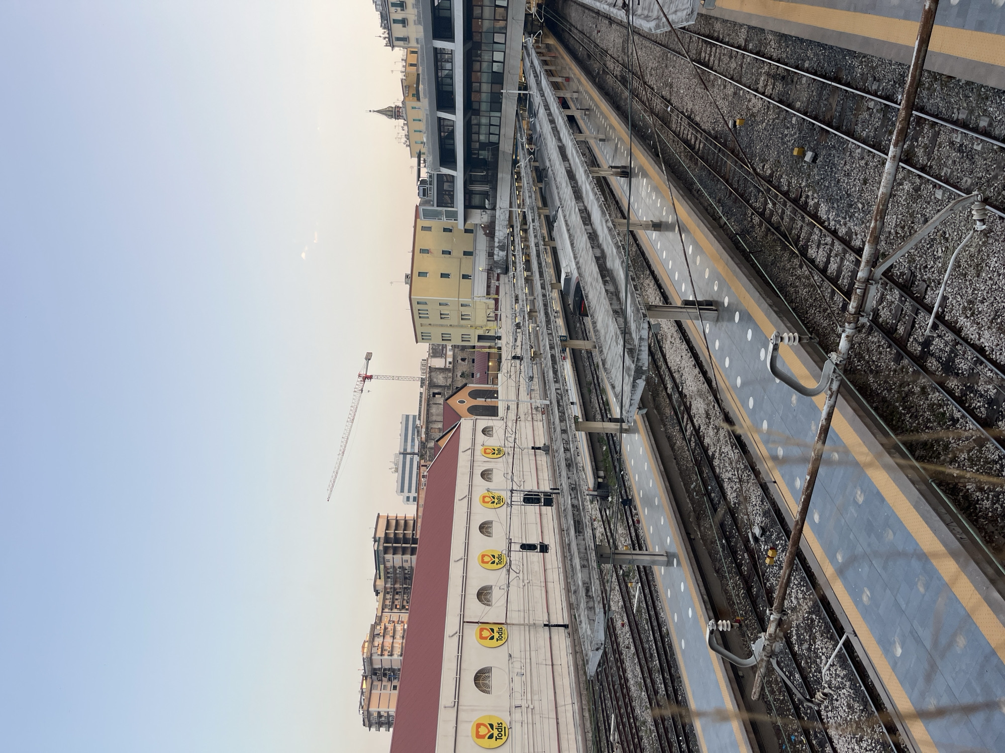 This is an eye-level, medium shot of a train station in Naples, Italy, likely taken during the late afternoon or early evening, given the warm lighting and clear sky. The foreground is dominated by train tracks and platforms. Several platforms are visible, featuring a modern design with blue and yellow markings, and circular patterns of white dots. Overhead power lines for the trains crisscross the scene, with some showing signs of rust. In the middle ground, a large, long building with a red roof stretches across the frame. This building has a white facade adorned with several circular yellow signs featuring a flower and the text "Todis." Behind this building, more structures are visible, including apartment buildings, a crane in the distance, and a church steeple, indicative of the urban environment of Naples. The scene appears to be a functional train station, with no people or trains immediately visible in the frame.