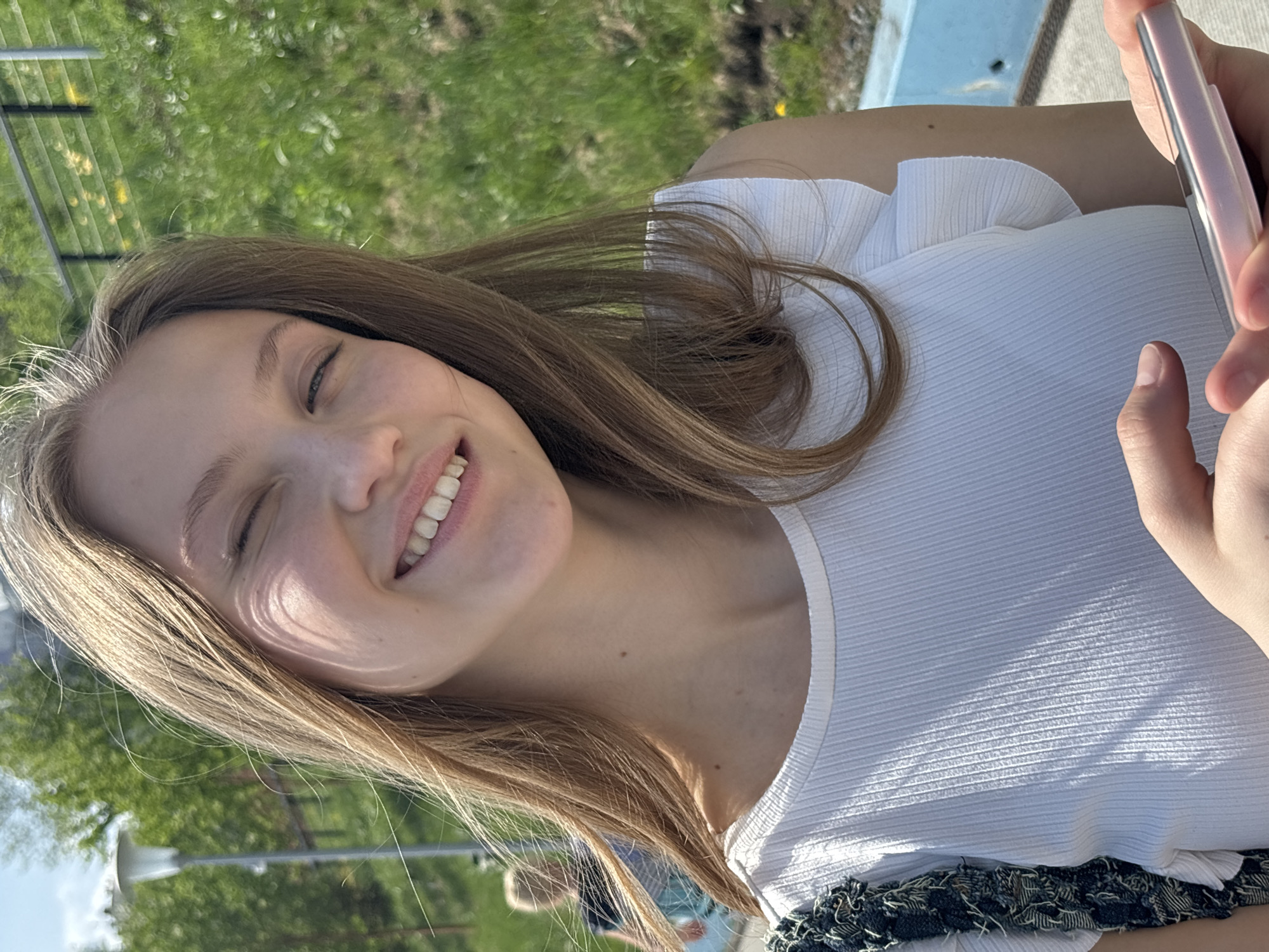 A young woman smiles while holding a pink cellphone in her right hand. She is wearing a white shirt. The background shows a green lawn, trees, and a fence, suggesting a park-like setting. A light post is visible. The image was taken outside in daylight. Location information is not deducible.