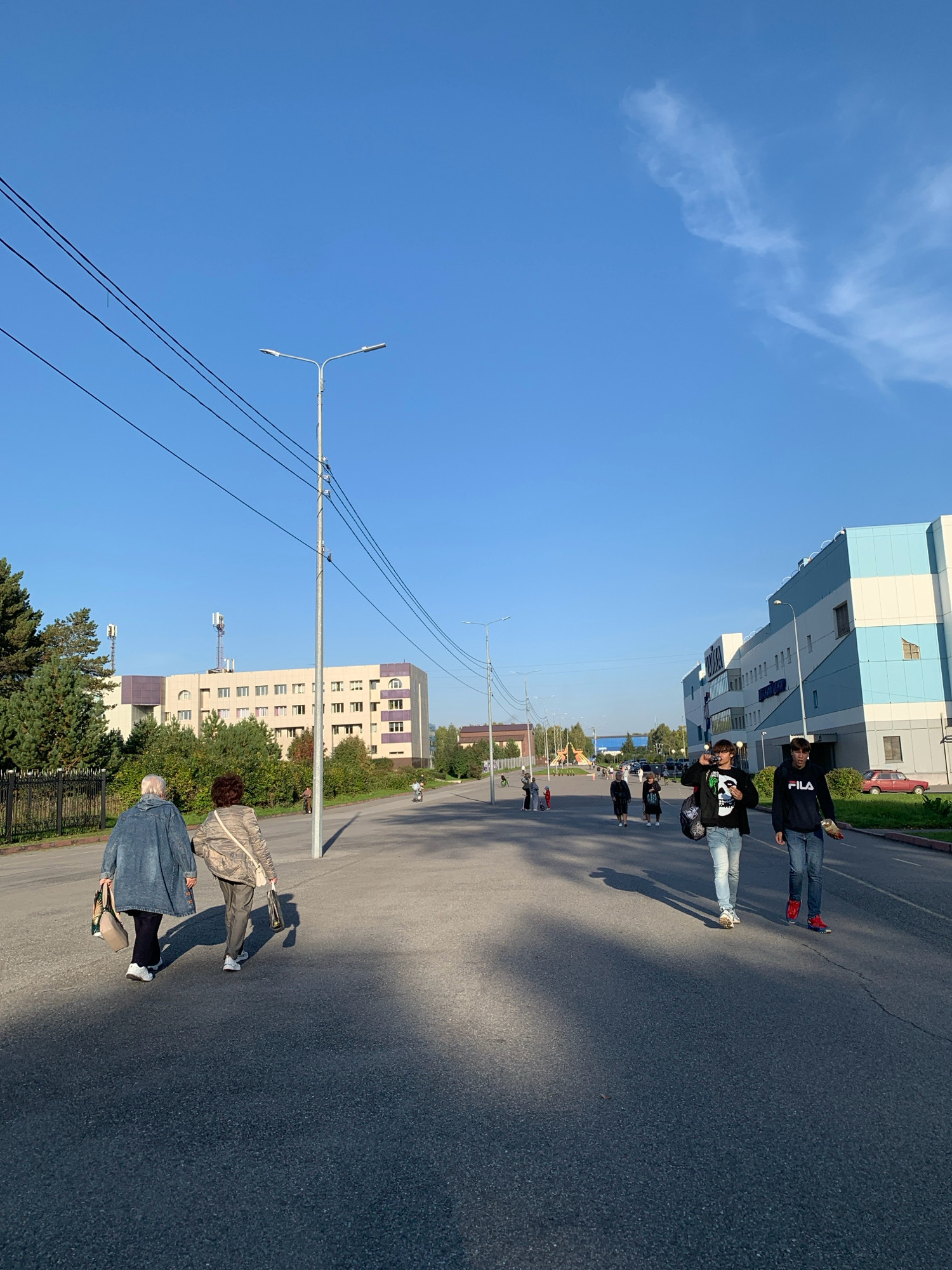 The image depicts a street scene in Prokopyevsk, Russia. In the foreground, two elderly women walk away from the camera, carrying shopping bags. They are walking on an asphalt road, with a shadow cast by a streetlamp on the right side of the frame. Further down the road, a group of younger individuals, two young men prominent in the center, walk towards the camera. One of the young men is drinking from a can. In the background, there are several buildings, including a modern, blue and white structure on the right and a light-colored building with purple accents on the left, along with trees and power lines. The sky is clear and blue with a wispy cloud formation. The overall impression is of a sunny day in a semi-urban environment.