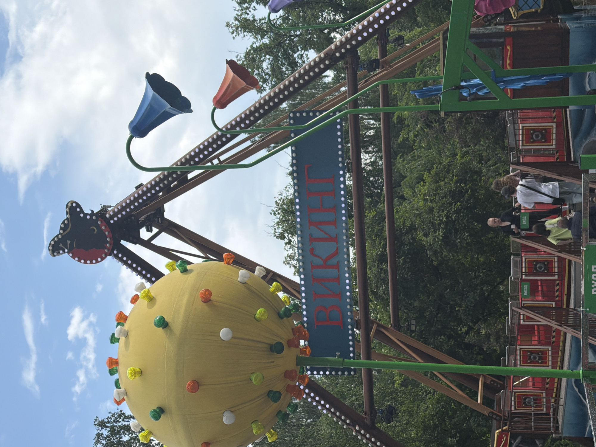 The image shows a brightly colored amusement park ride, possibly a pendulum ride, under a cloudy blue sky. A large yellow sphere, decorated with colorful lights, is positioned prominently in the foreground. To the right of the sphere, a large metal structure, part of the ride mechanism, extends upwards. This structure is adorned with rows of small white lights and features decorative bell-shaped elements in blue, red, and purple. A sign with red Cyrillic lettering, reading "ВИКИНГ" (Viking), is visible on a teal-colored board below the yellow sphere. In the background, lush green trees form a dense canopy, suggesting a natural or park-like setting. At the base of the ride, a red and gold passenger platform with railings is visible, and several people are standing or sitting on it. One person in the foreground, wearing a white shirt and a baseball cap, is looking away from the camera. Another person, with a shaved head and wearing a black top, is visible near the platform. The overall impression is of a lively, fun-filled atmosphere typical of an amusement park.