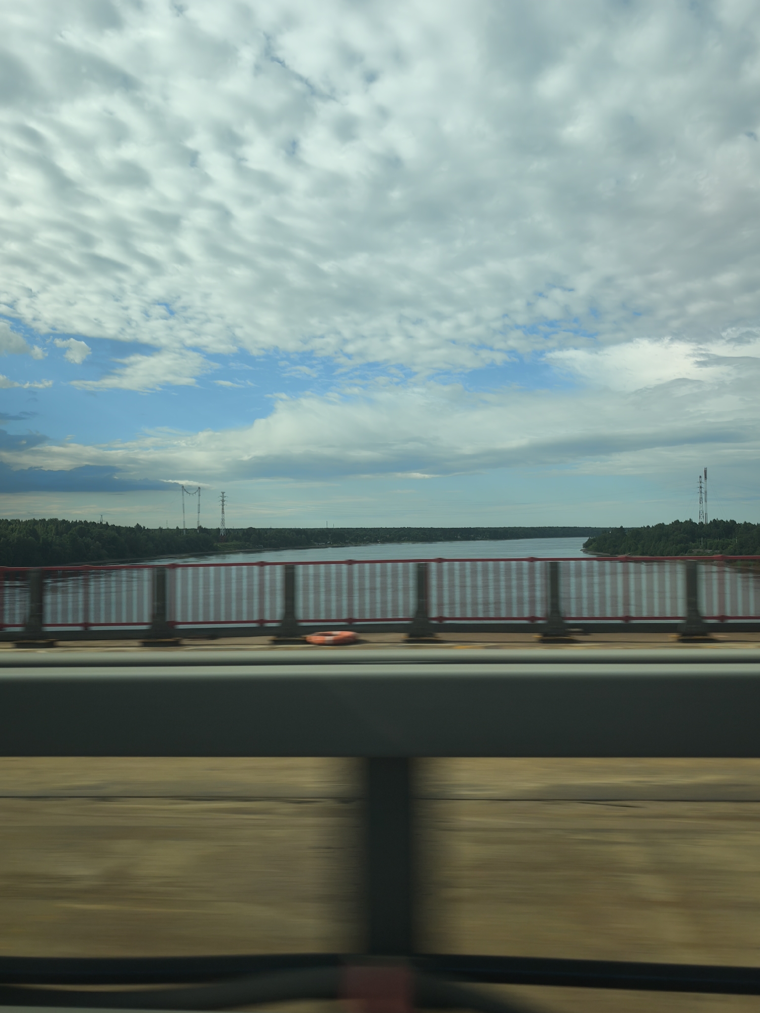 The image shows a view from a bridge overlooking a body of water, with trees and power lines on the opposite shore. The sky is partly cloudy. The bridge railing, painted red and white, is in the foreground. The overall scene suggests a transportation route within or near the city of Kirovsk, Russia. The movement blur indicates the photo was taken from a moving vehicle.