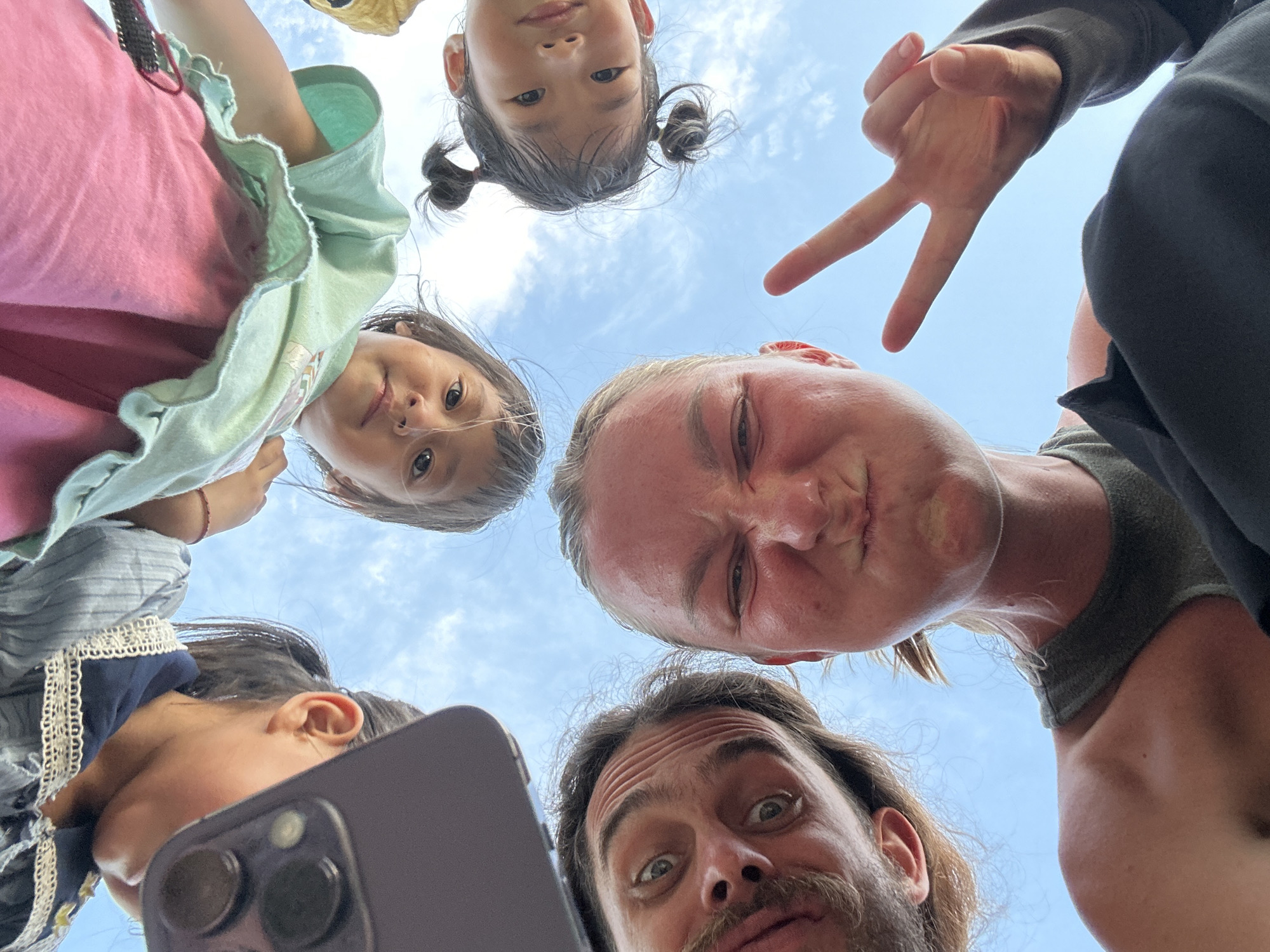 The image displays five individuals—two adult females, one adult male, and two young girls—looking downward into a camera against a background of clear blue sky with sparse, wispy white clouds. No specific urban or geographical features of Gia Hội, Vietnam, are visible within the frame, which predominantly features the sky.
In the upper-left quadrant, the top portion of an adult female's head, including her ear and part of her cheek, is partially obscured by a dark-colored smartphone held close to the lens. The phone's dual camera lenses are clearly visible. Above the center, a young girl with dark hair and a serious expression wears a light green ruffled top over a pink garment. To her right, in the upper-right quadrant, another young girl with dark hair styled in two small pigtails exhibits a gentle smile, wearing a yellow top.
At the lower-left, an adult male with a dark beard, mustache, and a visibly furrowed brow looks into the lens with wide eyes. Centered at the bottom and extending to the lower-right, an adult female with blonde hair and a tanned complexion makes an exaggerated, scrunched-up facial expression, with pouted lips and a squinted left eye. Her right hand is raised, forming a peace sign with her index and middle fingers extended. She wears a dark olive green tank top. The collective gaze and perspective indicate the photograph was taken from a low angle, with the subjects positioned directly above the camera.