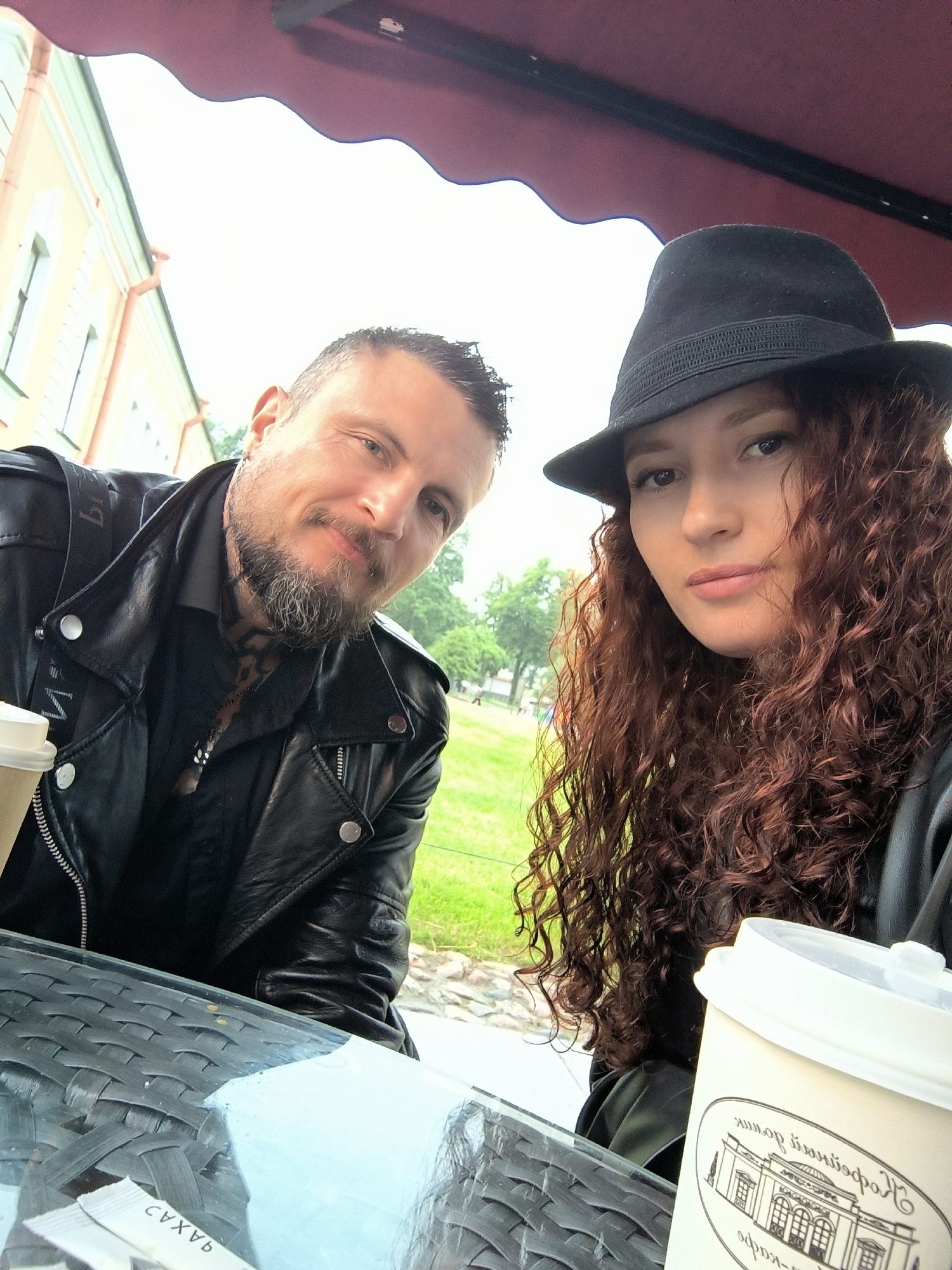 A man and a woman are taking a selfie at an outdoor cafe with a red awning. The man, with a beard and tattoos, is wearing a black leather jacket and a black shirt. The woman has long curly red hair and is wearing a black hat and a black jacket. They are sitting at a table, and there are two coffee cups on the table. In the background, there is a building with light-colored walls and green window frames, characteristic of Saint Petersburg, Russia. The scene appears to be a casual outing in the city.