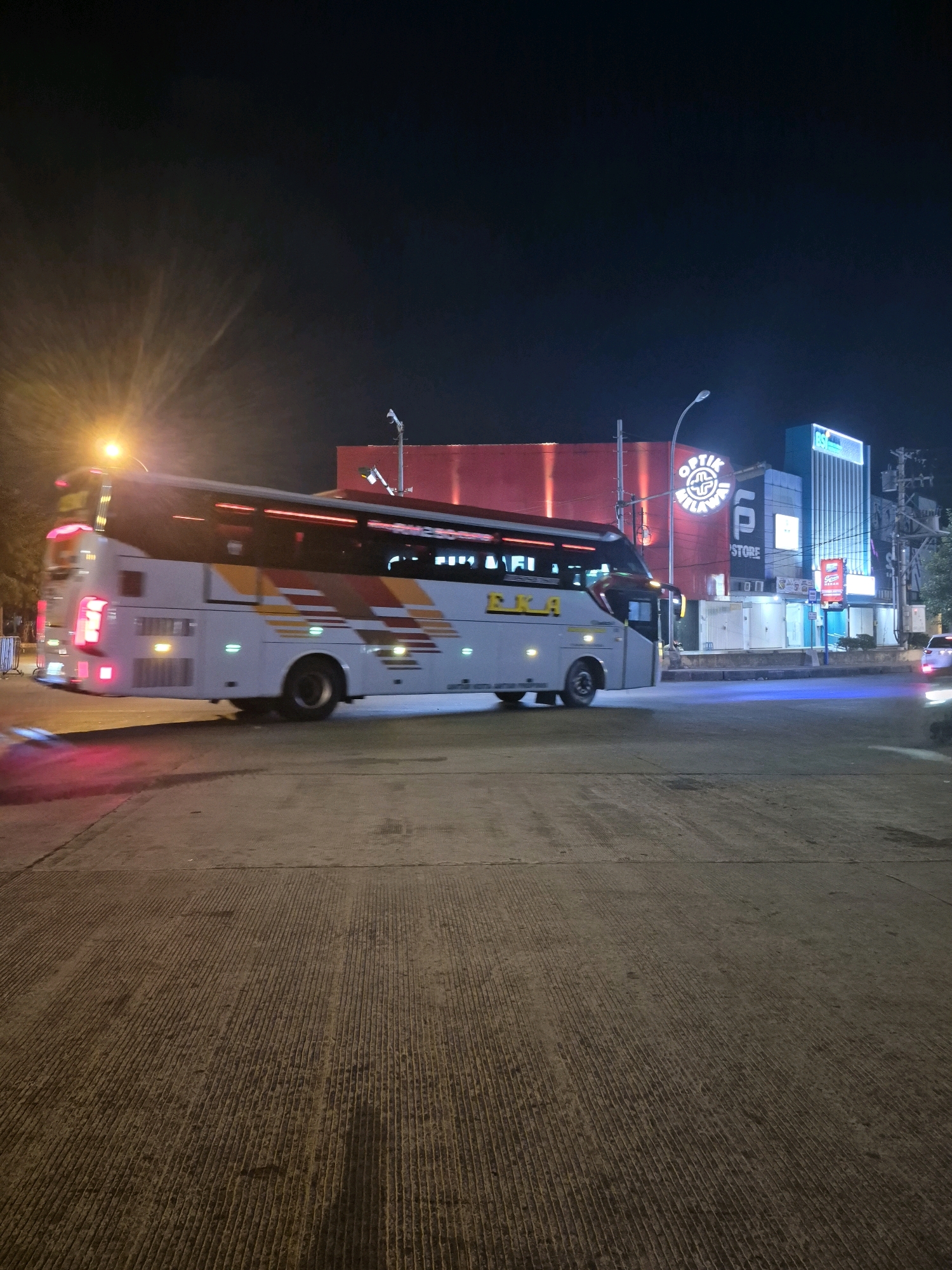 A white bus with red, yellow, and orange stripes, identified by the text "PKA" on its side, is captured in motion at night on a paved road in Surakarta, Indonesia. The bus is illuminated by its own lights and the surrounding neon signage of businesses. In the background, a large red building is visible, along with signs for "OPTICK GALAWAI" and "SP STORE." The scene suggests a commercial area with active businesses operating into the evening. The sky is dark, indicating it is nighttime, and the ground is a textured concrete surface.