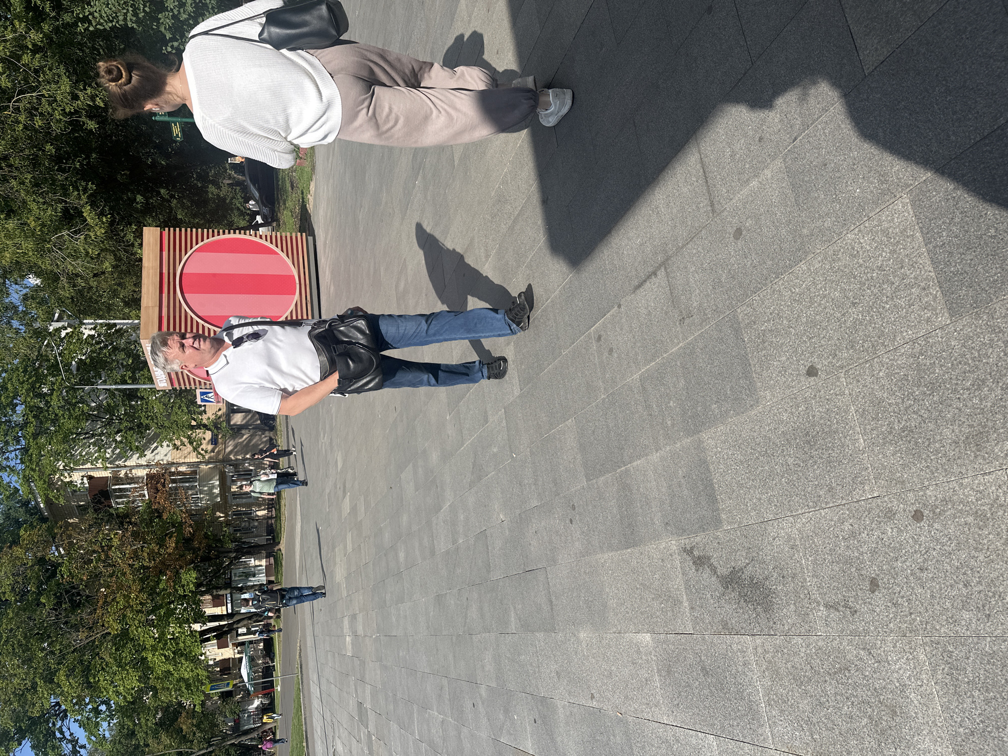 The image depicts a sunlit, paved plaza in Moscow, Russia, with several individuals walking. In the foreground, a man in a white polo shirt and blue jeans walks towards the left, carrying a black shoulder bag. To his right, a woman in a white sweater and light-colored pants walks away from the camera, also with a black shoulder bag. Behind them, a vibrant red and pink cylindrical structure stands. In the background, trees line the plaza, and apartment buildings are visible. Several other people are seen walking in the distance, and the ground is paved with large grey tiles. The scene suggests a typical urban environment on a bright day.