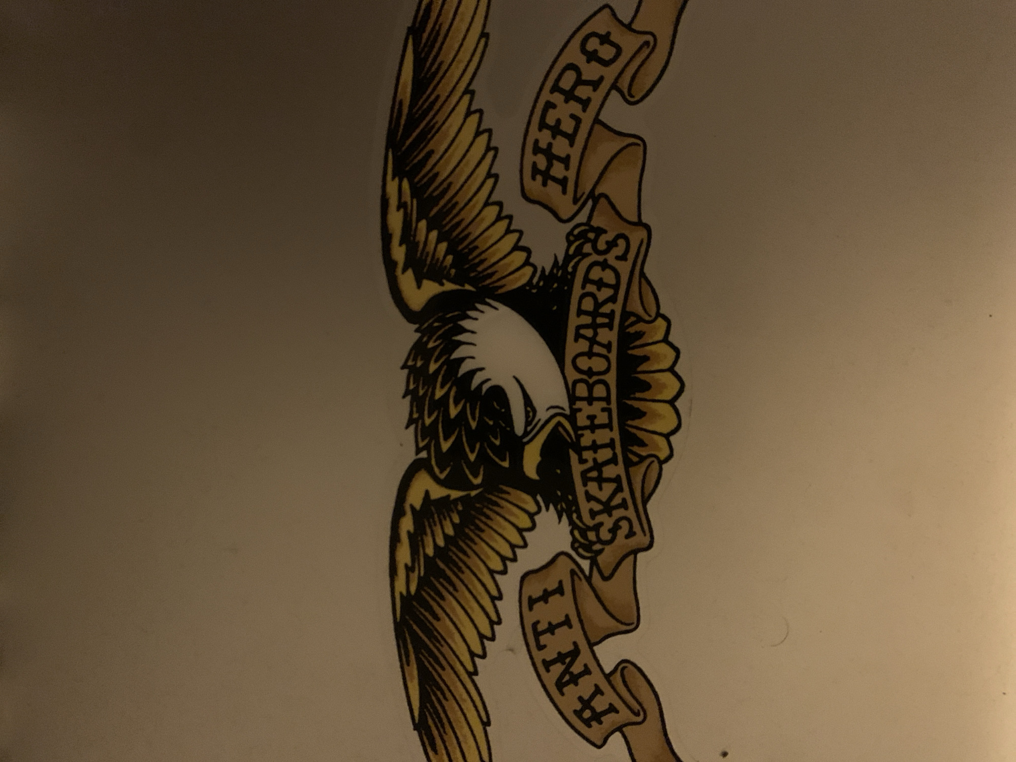 The image shows a stylized drawing of a bald eagle with outstretched wings, clutching a banner with the words "ANTI HERO SKATEBOARDS". The word "HERO" is above the eagle's head and "ANTI" is below. The drawing is predominantly in black and white with yellowish-tan highlights. The background appears to be a plain surface. The image shows the ANTI HERO Skateboards logo. There is no indication that this image is from Barcelona, Spain.