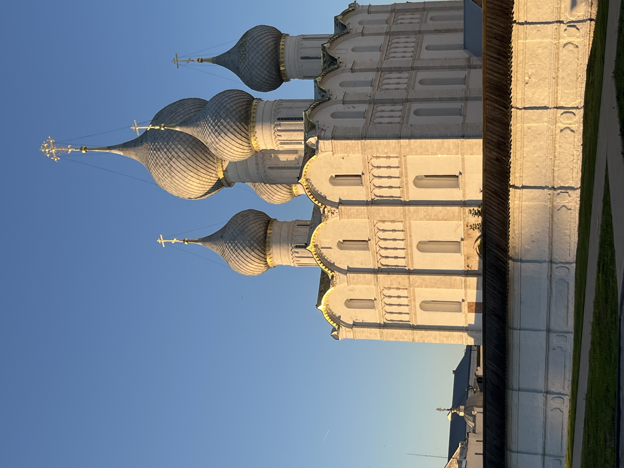 The image shows a large, white, Orthodox church with multiple domes, typical of Russian architecture. The church is bathed in the warm light of a setting sun, casting long shadows. The domes are silver-colored and topped with golden crosses. The building's facade features several arched windows and decorative elements. In the foreground, there is a white wall, likely part of the church's complex or a surrounding fortification, with a wooden roofline. A patch of green grass and a pathway are visible in front of the wall. The sky is a clear, bright blue, indicating a sunny day. The image appears to be taken from a slightly low angle, emphasizing the grandeur of the church. The location is identified as the city of Kladovitsy, Russia. There are no people visible in the image.