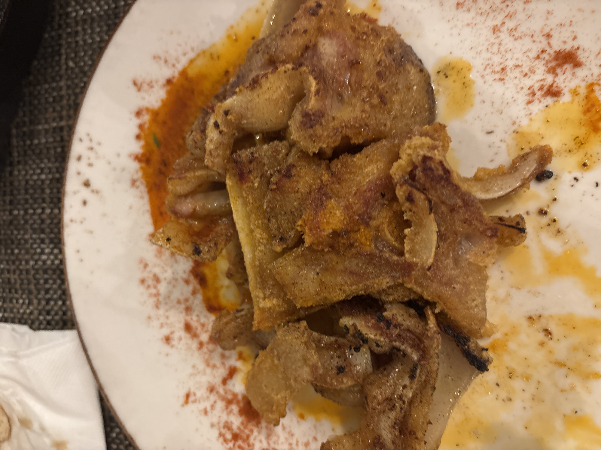 The image shows a plate of food. The food appears to be fried pork ears, cut into irregular shapes and piled on top of each other. The plate is white and has a scattering of red paprika powder and some oily sauce. The background is a dark, textured surface. There is no evidence to suggest that the location is Sofia, Bulgaria. There are no people in the scene.