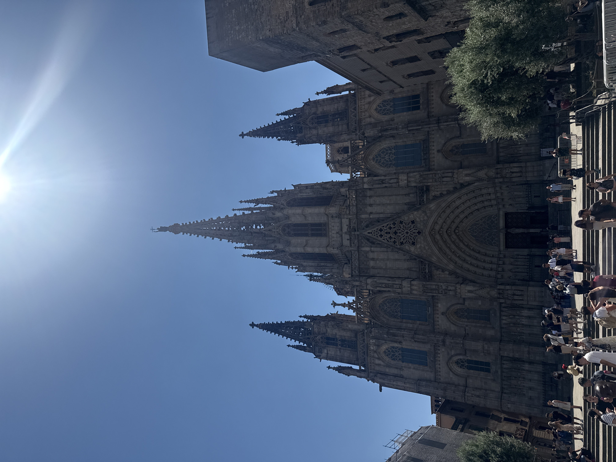 The image shows a wide-angle shot of the Barcelona Cathedral's exterior under a bright blue sky. The sun is high in the sky, indicating it is likely midday. The architecture of the cathedral is Gothic, with intricate stone carvings, pointed arches, and spires reaching towards the sky. The facade is a dark grey stone, showing signs of age and weathering. A large green tree is visible on the right side of the image, near the entrance.
A crowd of tourists is gathered on the steps leading up to the cathedral entrance. They are dressed in casual summer clothing, suggesting warm weather. Some people are taking photos, while others are milling about, looking at the architecture. The overall atmosphere appears to be one of tourism and sightseeing. The people are diverse, with no particular emotional expression evident in the crowd.
The image is taken from a low angle, emphasizing the grandeur and height of the cathedral. The perspective is also slightly tilted, giving a dynamic feel to the scene. The sun is shining intensely, creating a bright lens flare on the left side of the image, which adds to the sense of a sunny day. There is no visible text in the image. The setting is clearly a public space in Barcelona, Spain, given the architectural style of the cathedral and the presence of tourists.