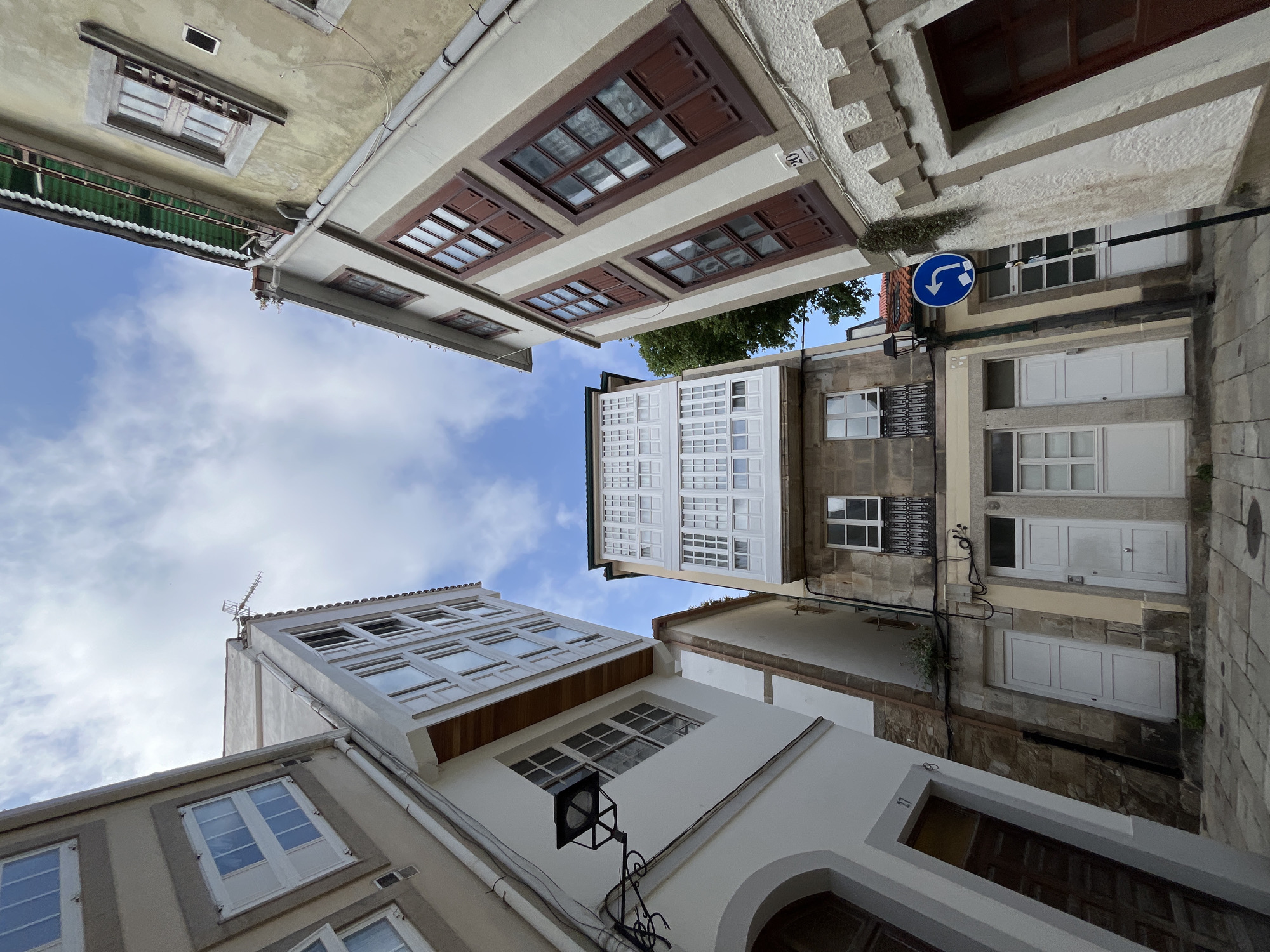 The image is taken from a low angle looking upwards in a narrow street in A Coruña, Spain. The buildings on either side of the street create a V-shape against the sky. The buildings are primarily cream and white, with windows of various sizes and styles. Some windows have dark wooden frames with divided panes, while others are white. One building features a white balcony with multiple panes. A blue and white directional sign is visible on one side of the street. The sky is visible in the space between the buildings, showing clouds. The street is narrow, with the buildings close together. There is no indication of people or other activity in the frame.