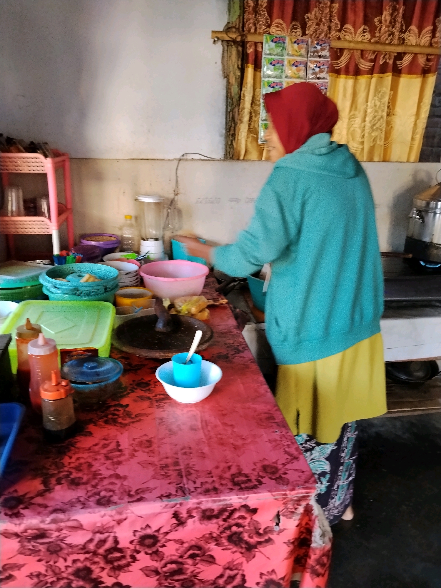 A woman wearing a red hijab and a teal hoodie is standing in what appears to be a kitchen or food stall in Panggungan, Indonesia. She is facing away from the camera, towards a stove where a pot is being heated by a blue flame. The kitchen counter is covered with a red floral tablecloth and is cluttered with various kitchen items, including bowls, utensils, and bottles of condiments. To the left of the woman is a pink plastic shelf unit with glasses and other items. Behind her, a window is covered by patterned curtains. The walls are a light color.