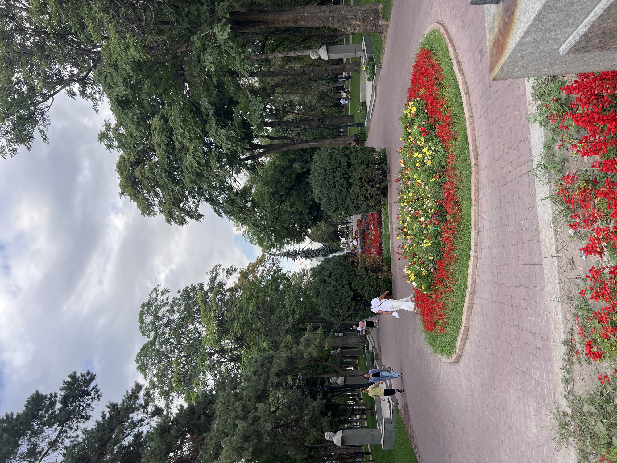 This is a medium shot of a park pathway in Bishkek, Kyrgyzstan. The pathway is paved with red bricks and curves to the right. On the left side of the pathway, there are several busts on pedestals, and trees. On the right side of the pathway, there is a large flower bed with red, yellow, and white flowers. There are several people walking on the pathway. The sky is cloudy. In the background, there is a fountain and more trees.