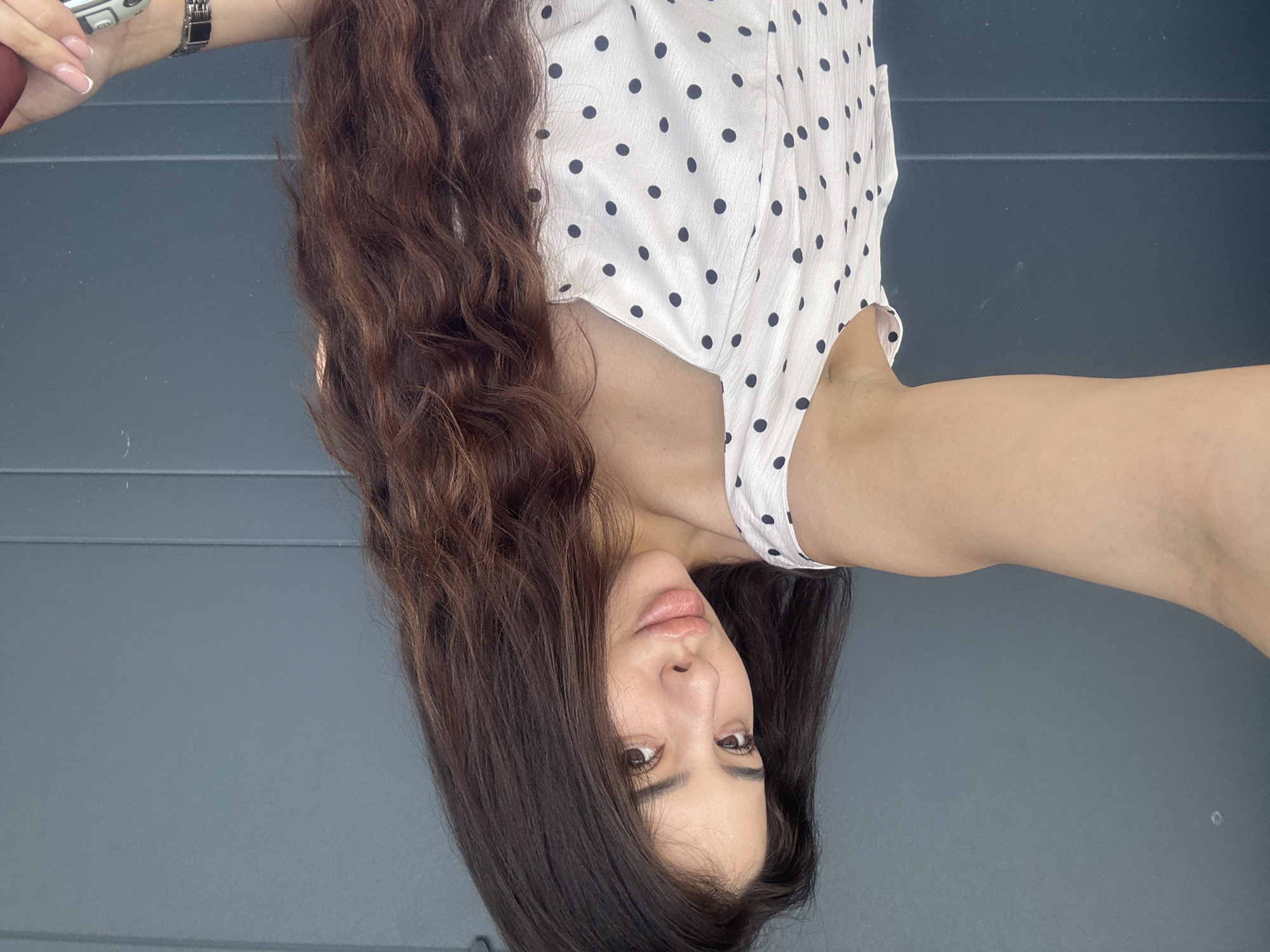 A woman is oriented upside down in the frame, wearing a white shirt with black polka dots. She has long, wavy brown hair. The background is a gray, textured wall. A silver watch is visible on her wrist. The location is not evident from the image, and there is no information to suggest the location is Plovdiv, Bulgaria.