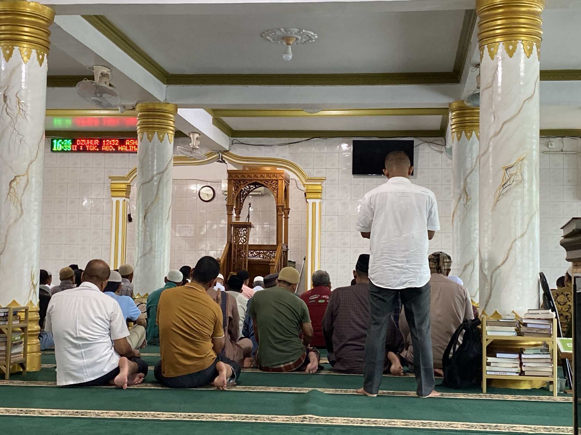 A group of Muslim men are gathered inside a mosque in Lhokseumawe, Indonesia, captured from a low-angle, rear perspective. Most men are seated on a green carpet with patterned borders, facing towards the front where a wooden minbar (pulpit) stands. One man in a white shirt and dark trousers is standing in the foreground, also facing forward, his back to the viewer. Several other men are sitting further back, some wearing traditional prayer caps (kopiah).
The mosque's interior features large white pillars adorned with golden capitals and a marble-like veining pattern. The ceiling is white with golden trim along the edges of beams. Two ceiling fans are visible, along with a decorative light fixture. The walls are tiled in a white pattern. A digital display board hangs on the wall between two pillars, showing information in green and red LEDs. To the right, a flat-screen television is mounted on the wall, and a traditional wall clock is visible further back near the minbar. A small wooden bookshelf filled with books is positioned to the right of the foreground, beside one of the pillars. A black backpack rests on the floor next to a seated man.
The digital display clearly indicates the current time as 16:35. It also shows prayer times: "DZUHUR 12:32" (Dhuhr) and "ASHAR 15:53" (Asr). A lower line of text reads "II : TGK. ABD. HALIM", likely indicating the name of a speaker, imam, or the mosque itself. The presence of the minbar and prayer times suggests a religious service, sermon, or congregational prayer is ongoing or about to commence, consistent with a mosque setting in Lhokseumawe. The atmosphere appears to be one of quiet reverence.