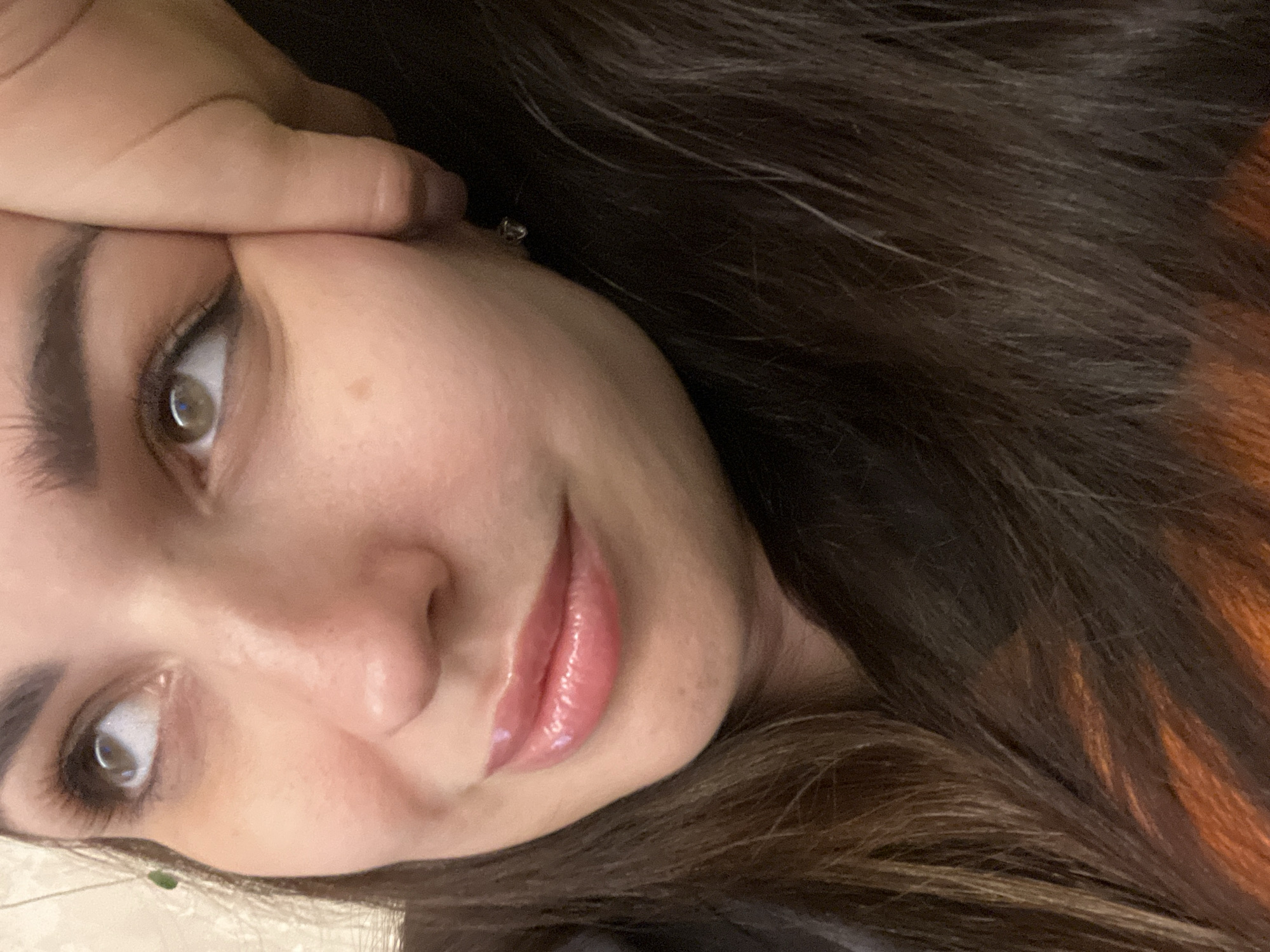 The image is a close-up, low-angle shot of a woman's face, with her right hand resting on her cheek and her head tilted upwards. She has dark brown hair, full lips with lip gloss, and is looking to her left. A small, clear earring is visible on her right earlobe. The background is out of focus, appearing to be a textured surface. The lighting is soft and warm, highlighting her skin.