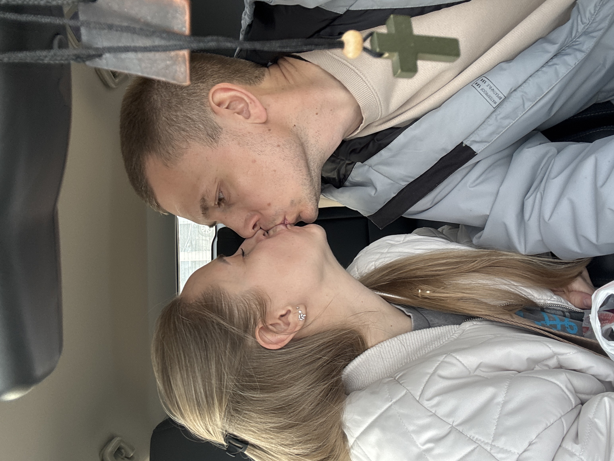 This intimate image, taken in Chaikovsky, Russia, captures a tender moment between a man and a woman sharing a kiss. The photo is rotated, showing the subjects as if lying on their sides, suggesting it was taken in a confined space, likely the interior of a vehicle.
The man, positioned on the left, has short, light brown hair and light stubble. He is dressed in a light beige long-sleeved shirt layered under a grey and black jacket. A small label is visible on his jacket sleeve, displaying the text "WATERPROOF 15K" and "BREATHABLE 15K."
The woman, on the right, has long, straight blonde hair. She is wearing a white quilted jacket over a grey top. Her left ear is adorned with two small silver earrings: a simple stud and a more elaborate floral-shaped piece. Their lips are gently touching, indicating a moment of affection and connection.
In the immediate foreground, slightly out of focus, a dark green cross pendant hangs from a black cord, accompanied by a small wooden bead. The background consists of blurred interior elements of the vehicle, including what appears to be a window or a bright light source. A partially visible plastic bag, with obscured blue text, can be seen near the woman's shoulder. The overall lighting suggests it is daytime. The image conveys a sense of warmth and closeness between the couple.