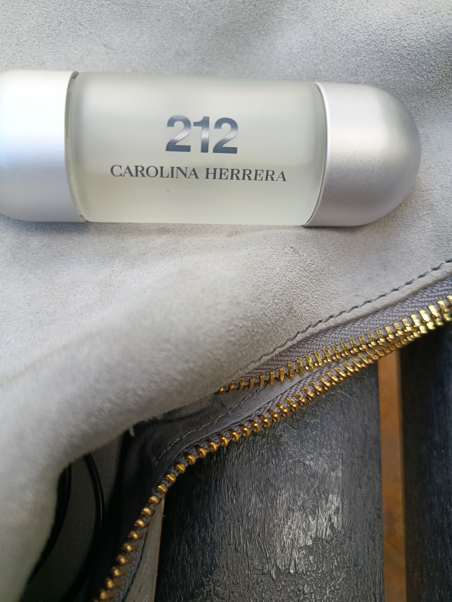 A horizontal, close-up, eye-level shot shows a bottle of Carolina Herrera 212 perfume resting on a light gray, fuzzy surface, possibly suede. The bottle is frosted white with silver end caps. The text "212" is prominently displayed in a dark gray font, with "CAROLINA HERRERA" printed below it in a smaller, darker font. A gold zipper with dark gray fabric is visible in the foreground, running diagonally across the bottom of the image. The background consists of a dark, textured surface, possibly leather, with a vertical, lighter brown object to the right. The scene is devoid of human presence and activity. The location is not discernible from the image.