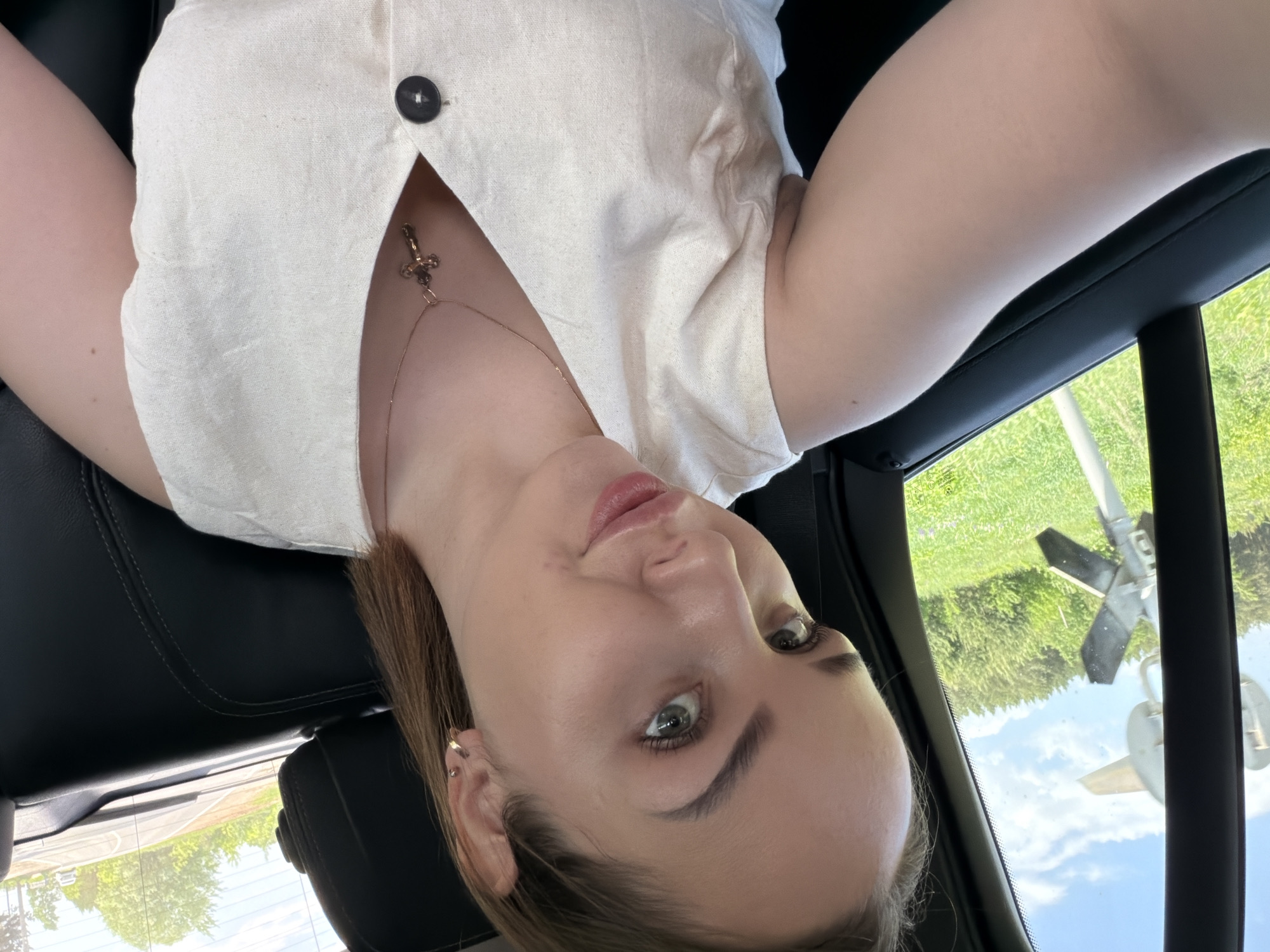 A woman takes a selfie from the back seat of a car. She has fair skin, green eyes, and light brown hair. She wears a light-colored button-down shirt and a gold necklace with a cross pendant. The scene outside the car window shows a railroad crossing signal. The exact location in the city of Chaikovsky, Russia cannot be determined.
