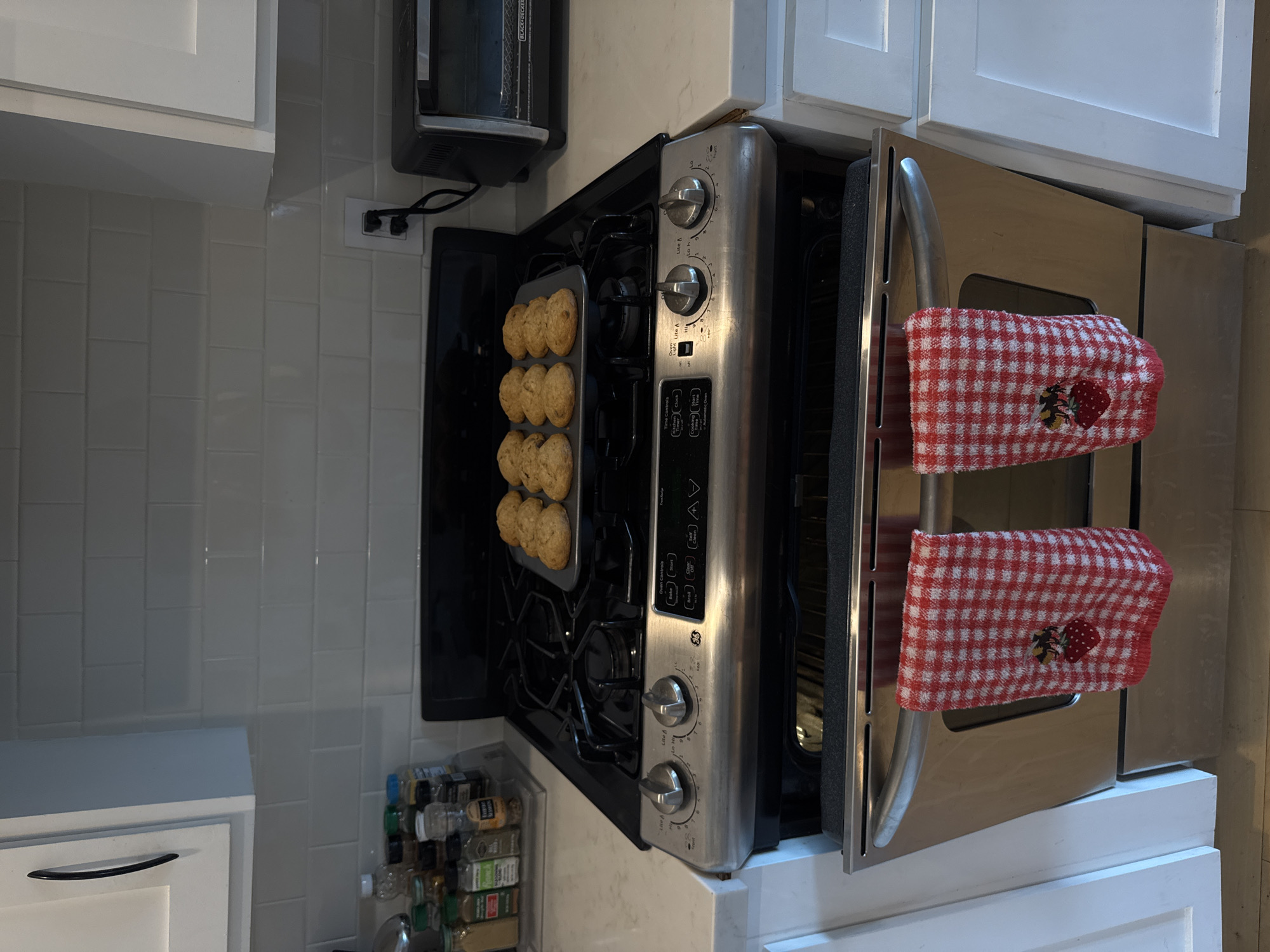 This image captures a domestic kitchen scene in Los Angeles, USA, centered around a stainless steel range and oven. The main activity is baking, with a metal baking tray containing nine light brown, freshly baked goods, likely muffins or scones, resting on the stovetop grates. The oven door is open, revealing the dark interior.
Draped over the oven's handle are two red and white checkered kitchen towels, each adorned with a strawberry and bumblebee design. The range itself features control knobs for the burners and a digital display panel with various function buttons. Visible text on the appliance includes "GE," "Oven Controls," "Time Controls," "Light," "Lite A," "Automatic Oven," along with labels like "Bake," "Broil," "Start," "Clock," and "Timer." Numerical settings for the burners are also visible around the knobs, ranging from "Lo" to "Hi."
The kitchen features white subway tile backsplash and white cabinets. To the upper left, a black "BLACK+DECKER" toaster oven or similar appliance is visible, plugged into a white electrical outlet. Below the range, on the left, a multi-tiered spice rack displays numerous transparent spice bottles. The scene is well-lit, typical of indoor daytime or evening lighting. No people are visible.