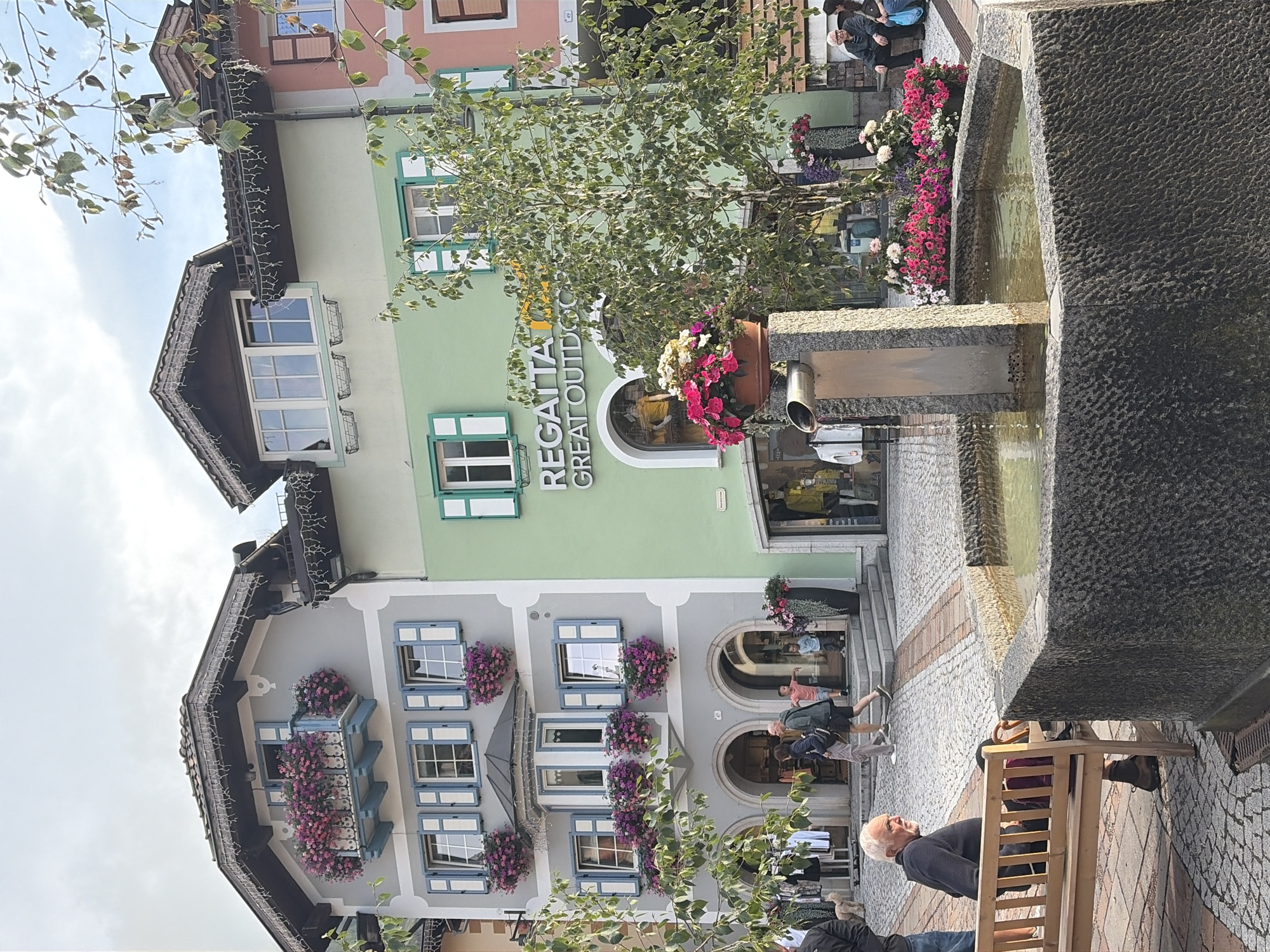 The image depicts a street scene in Moena, Italy, featuring a stone fountain in the foreground with water flowing from a metal spout. In the background, traditional Alpine-style buildings with colorful facades are visible. One building is light green and displays the "REGATTA GREAT OUTDOOR" signage. Adjacent to it is a gray building with blue shutters and numerous window boxes overflowing with pink flowers. People are scattered throughout the scene. An elderly man with white hair sits on a wooden bench to the left, facing away from the fountain. Further back, several individuals are walking on a cobblestone path, including a couple and two children. The overall environment is an outdoor public space, likely a town square or main street, characterized by pedestrian activity and architectural charm.